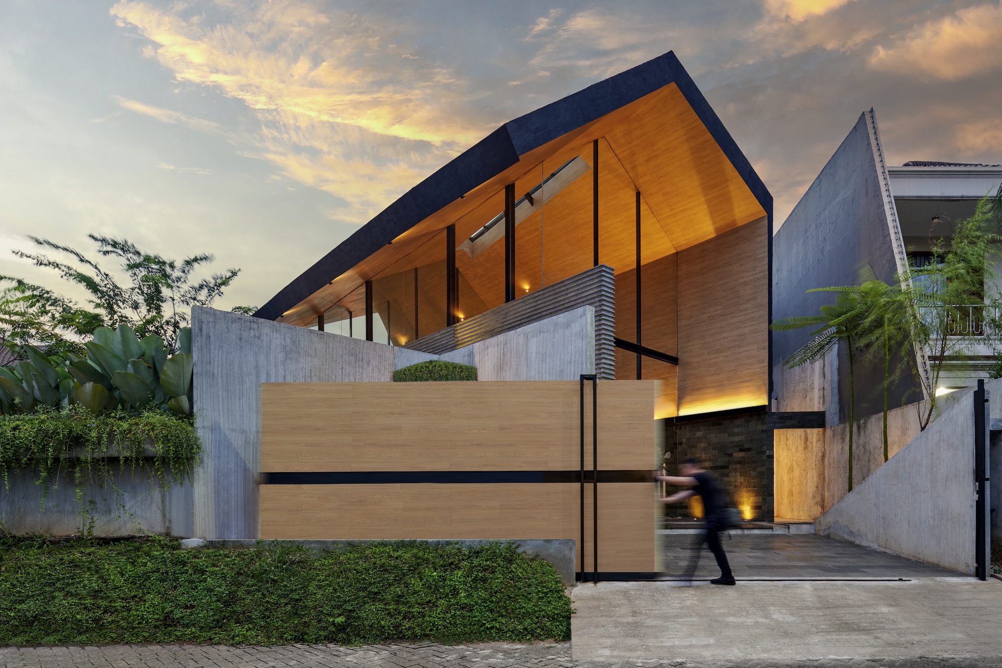 Street view of the timber-clad entrance with pivoting gate and figure walking into the courtyard at dusk