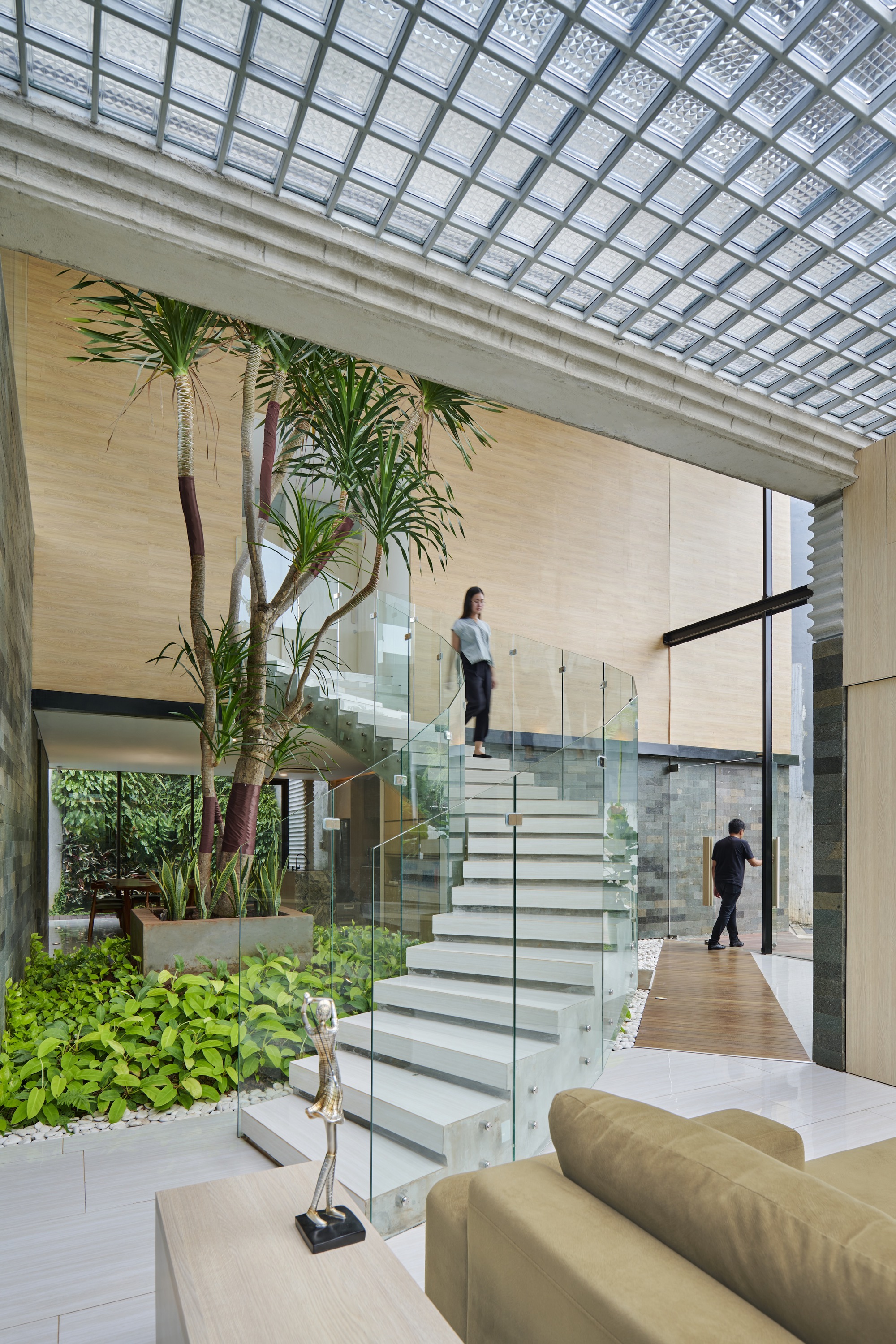 White staircase with glass railings ascending through a double-height atrium with gridded skylight and planted beds