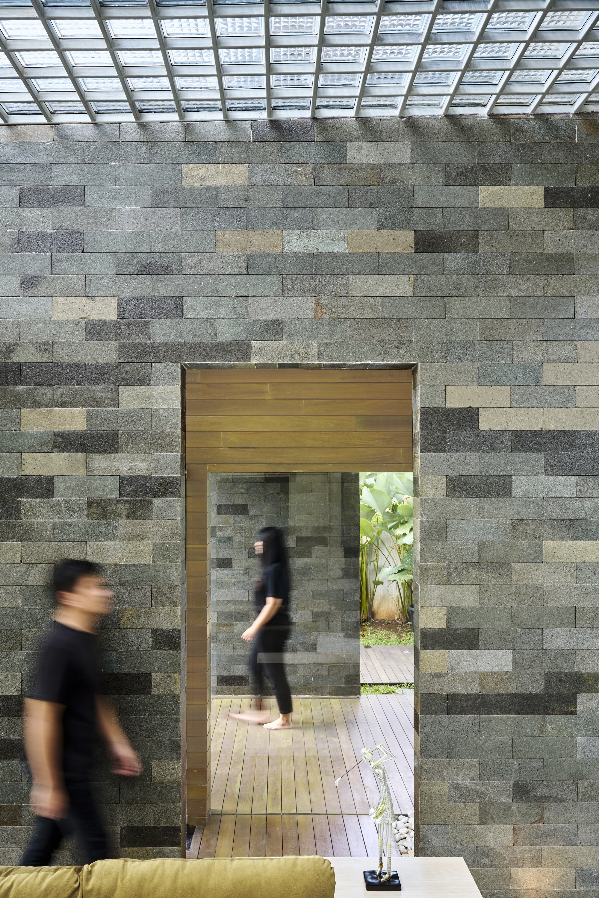 Timber-framed doorway through a variegated stone block wall with people passing beneath a slatted skylight