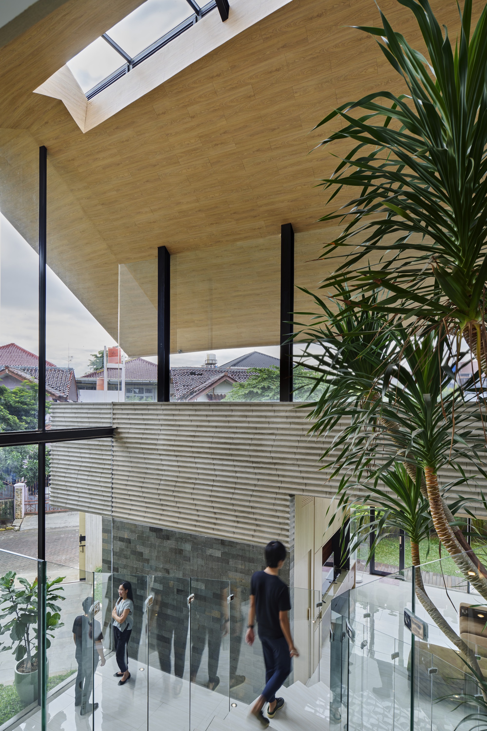 Double-height interior with plywood ceiling, black steel columns, glass balustrade and potted palms beside person descending
