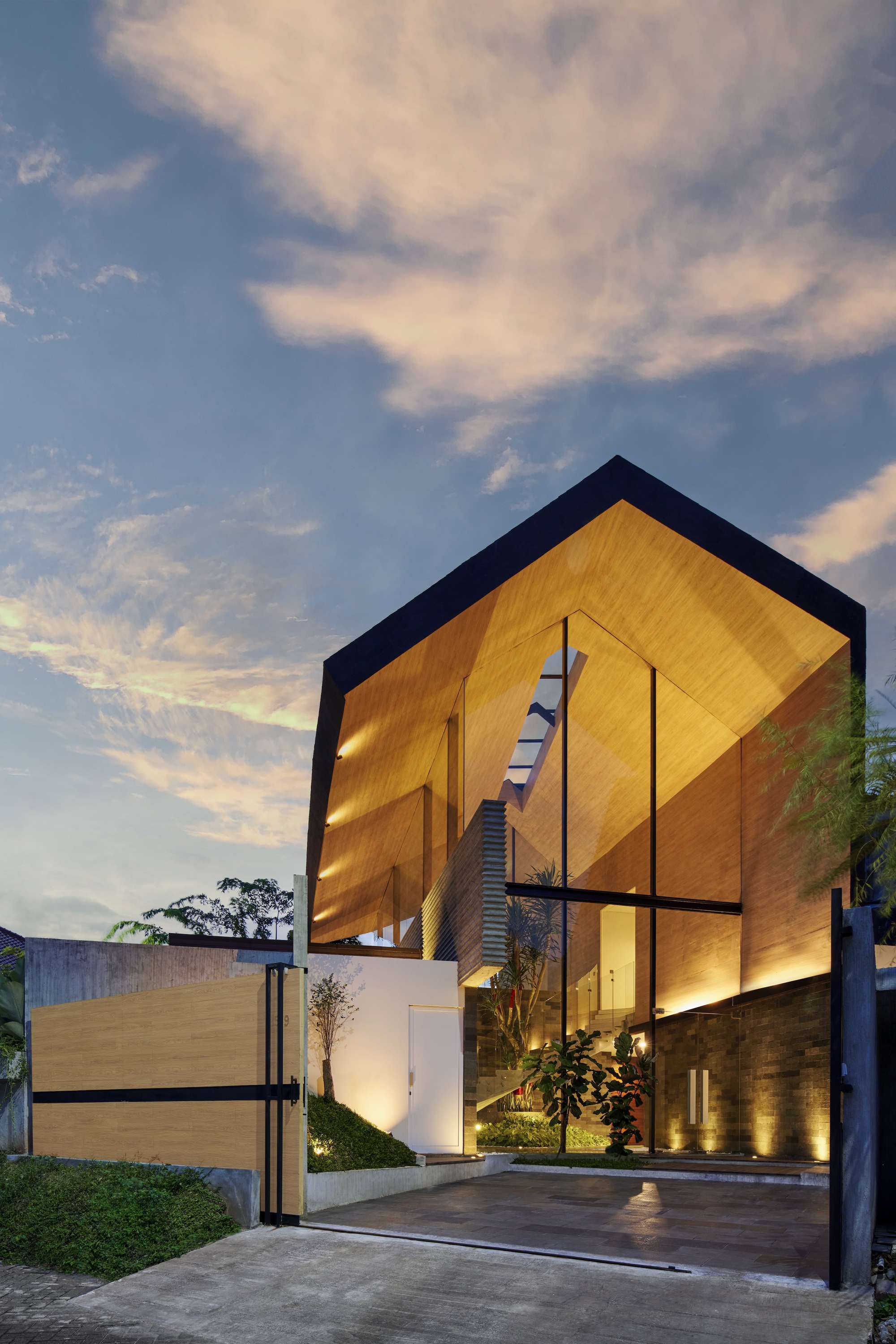Street view of the timber-clad gabled volume illuminated at dusk with a sliding gate entry