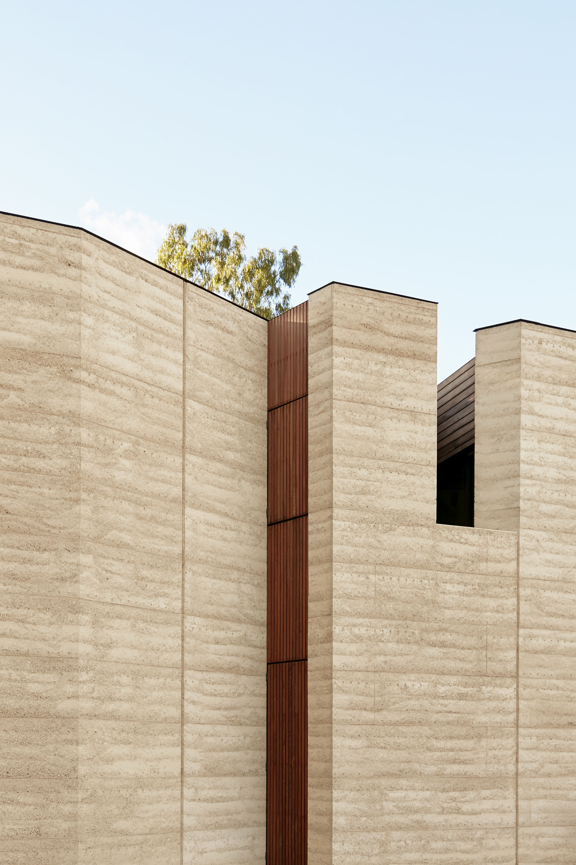 Rammed earth facade with vertical timber slot detail beneath a pale sky