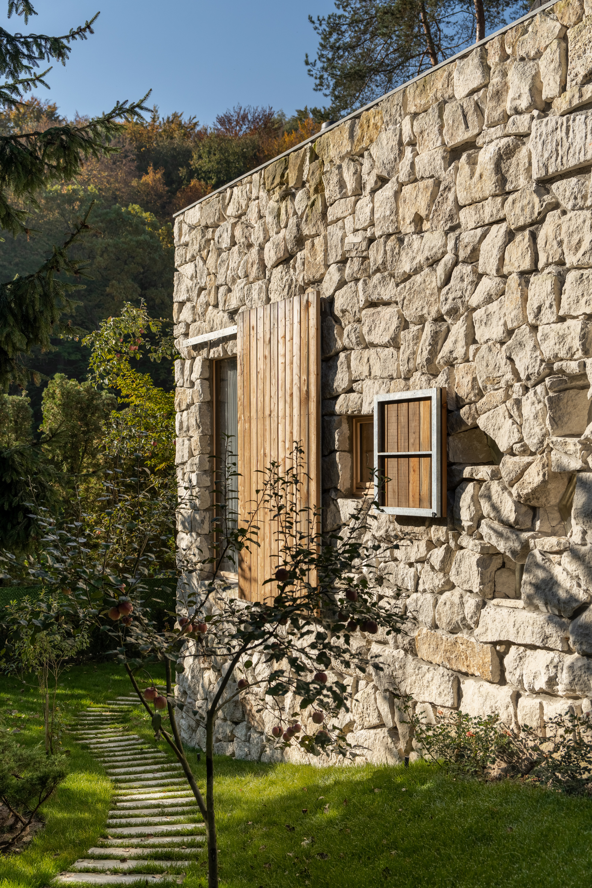 Rough stone wall punctuated by vertical timber cladding and window opening beside stepping stone path