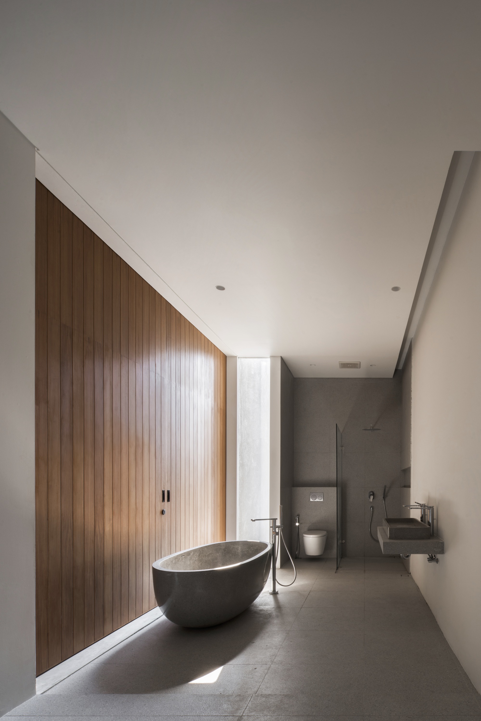 Long bathroom with freestanding stone bathtub beneath vertical timber paneling and clerestory window