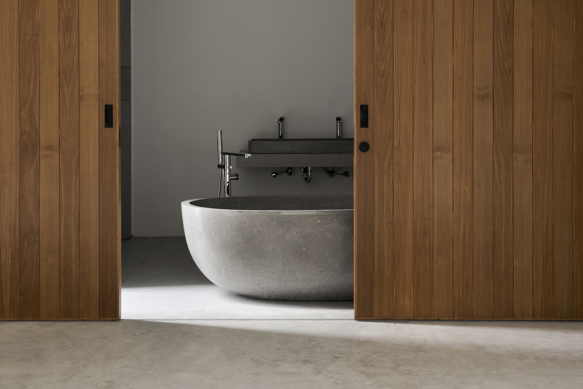 Freestanding stone bathtub framed by timber sliding doors with soft daylight across polished concrete floor