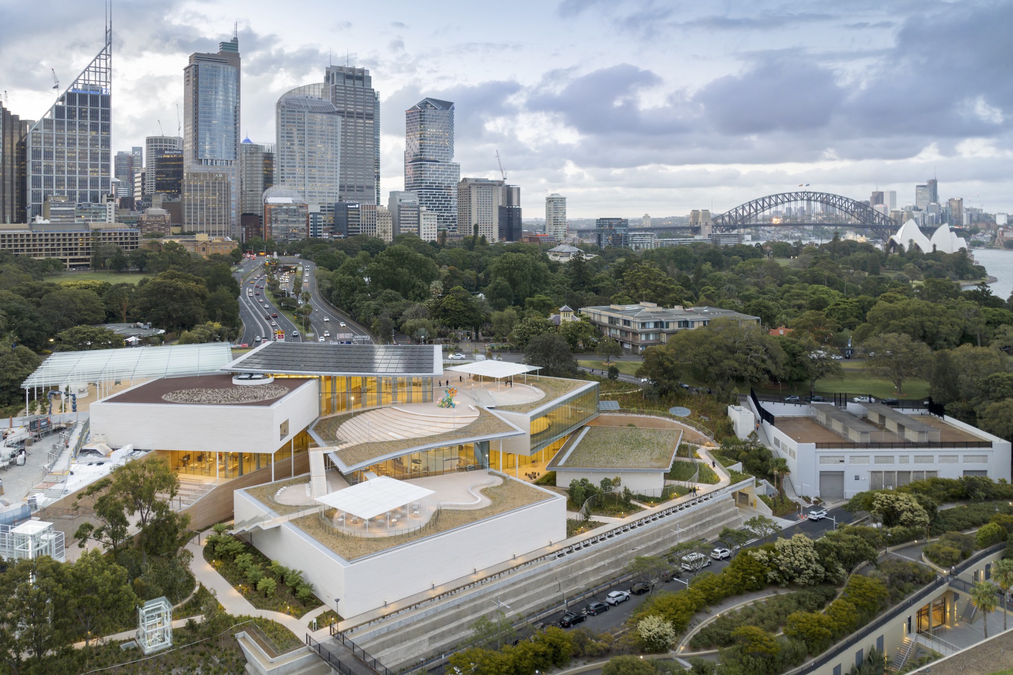 Aerial view of the terraced white volumes with golden glass walls nestled among trees and parkland