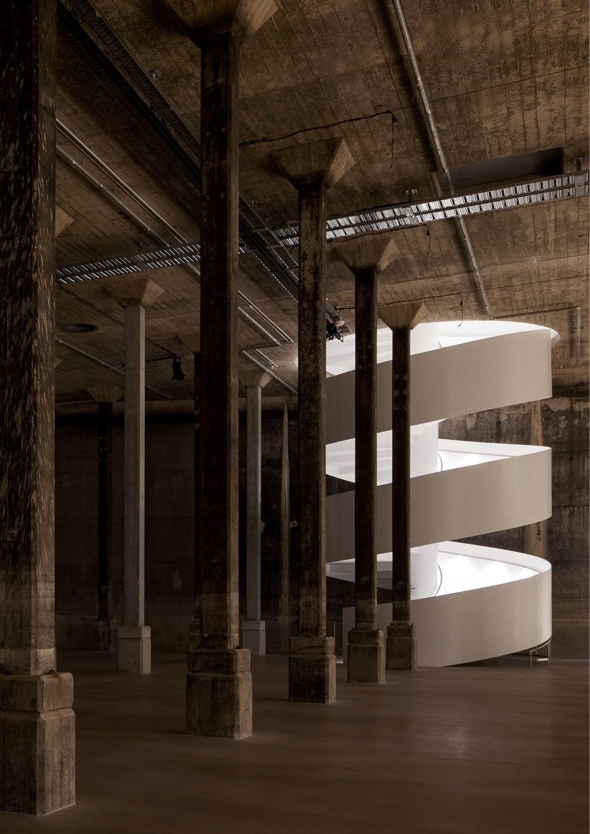White helical staircase illuminated by clerestory slot within a raw concrete hall with timber columns
