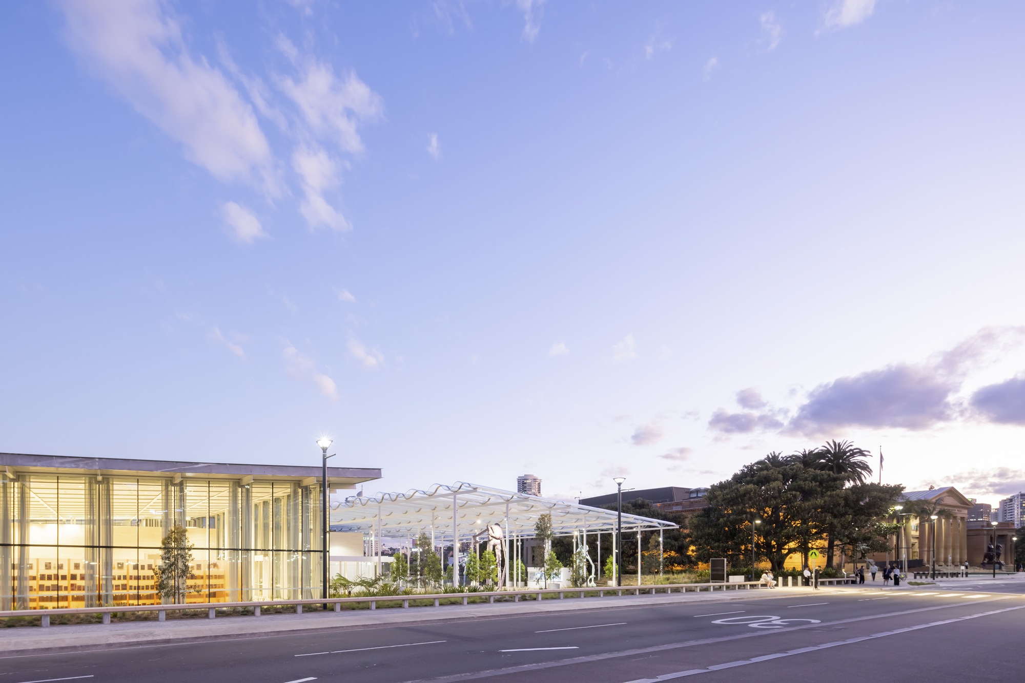 Street view of the glass-walled entrance pavilion and lattice roof structure at dusk with palm trees visible beyond