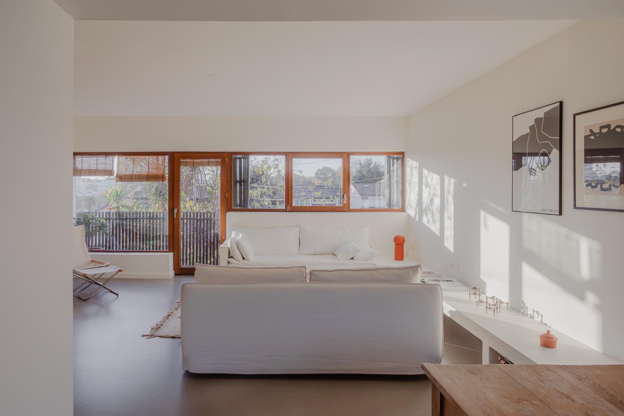 Living room with timber-framed ribbon windows overlooking a slatted fence and winter garden with strong afternoon shadows