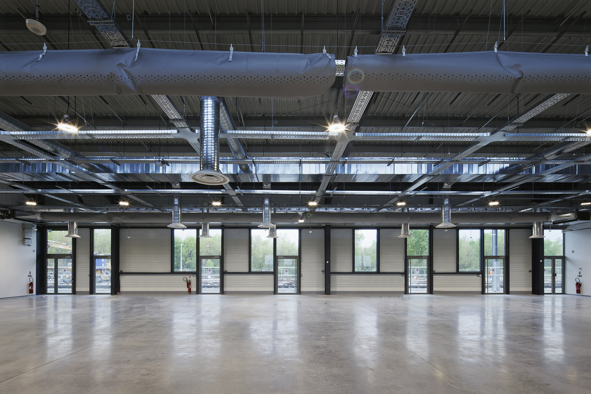Open-plan interior space with exposed ceiling ducts, pendant lights, and a glazed facade overlooking greenery