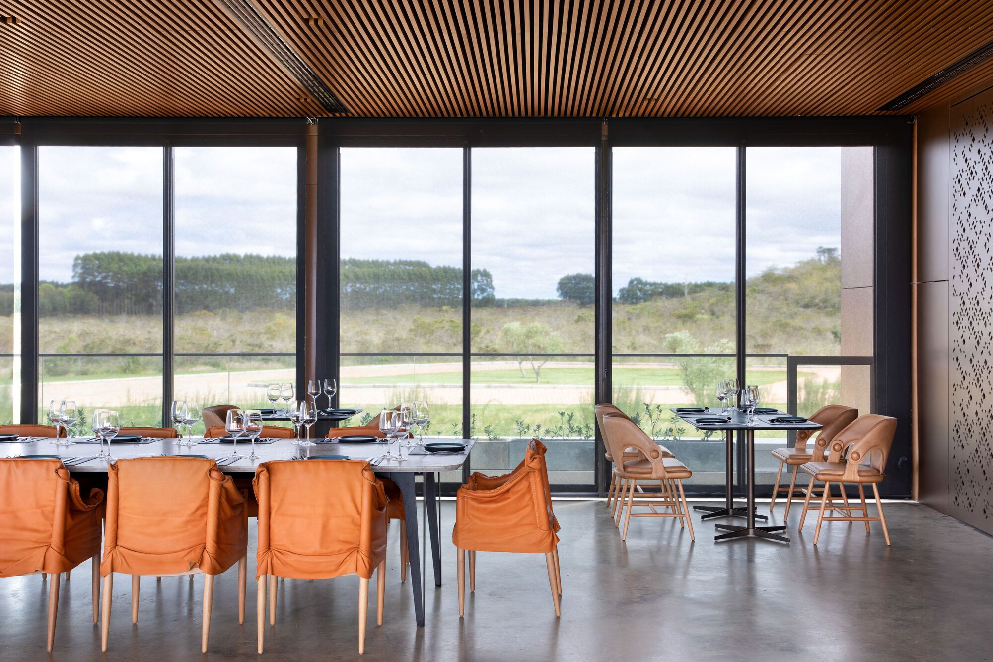 Dining space with orange upholstered chairs and slatted timber ceiling facing expansive glazing and landscape beyond