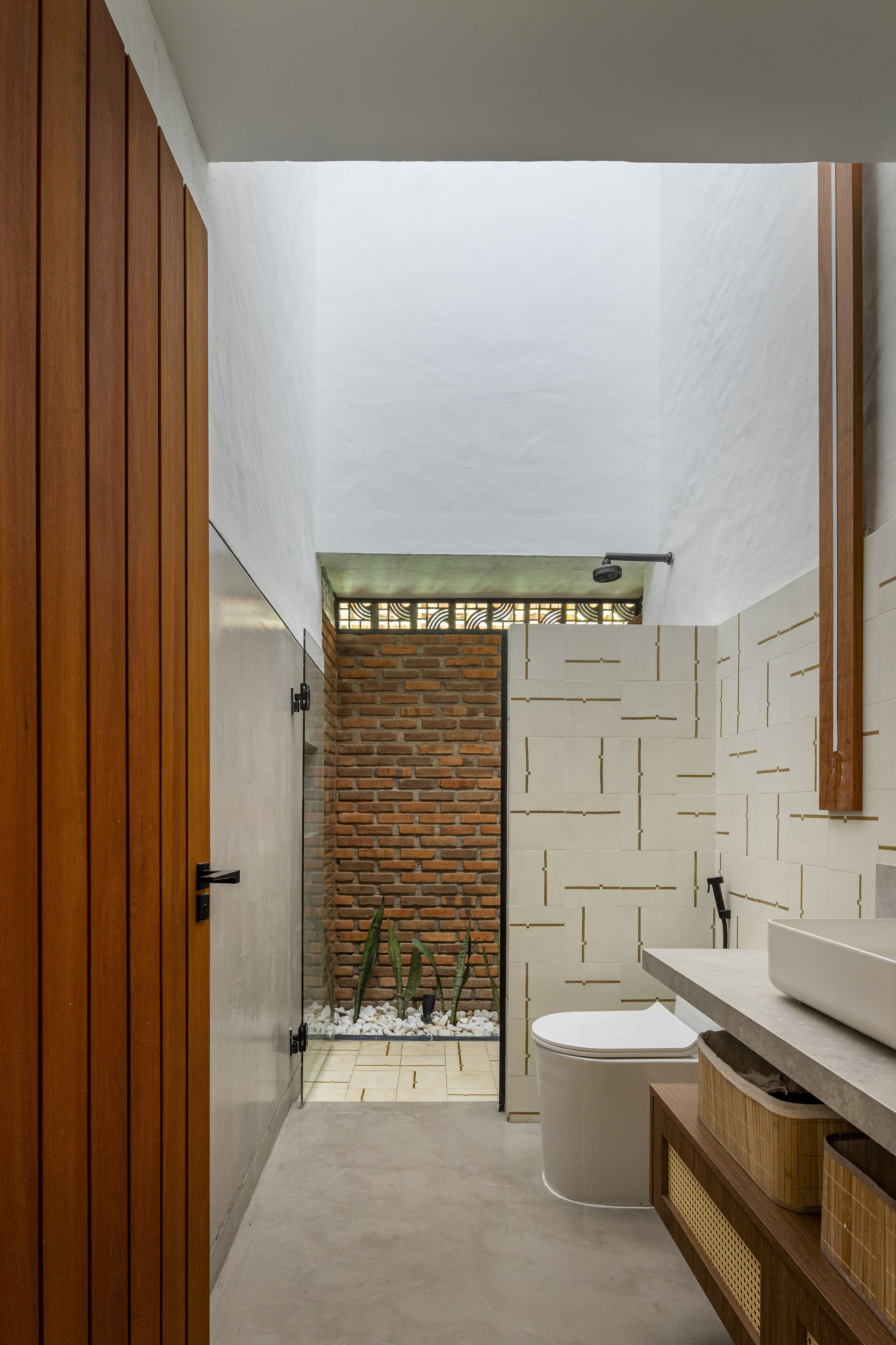 Narrow bathroom with tall ceiling and glass door opening to a brick-walled courtyard with planted gravel bed