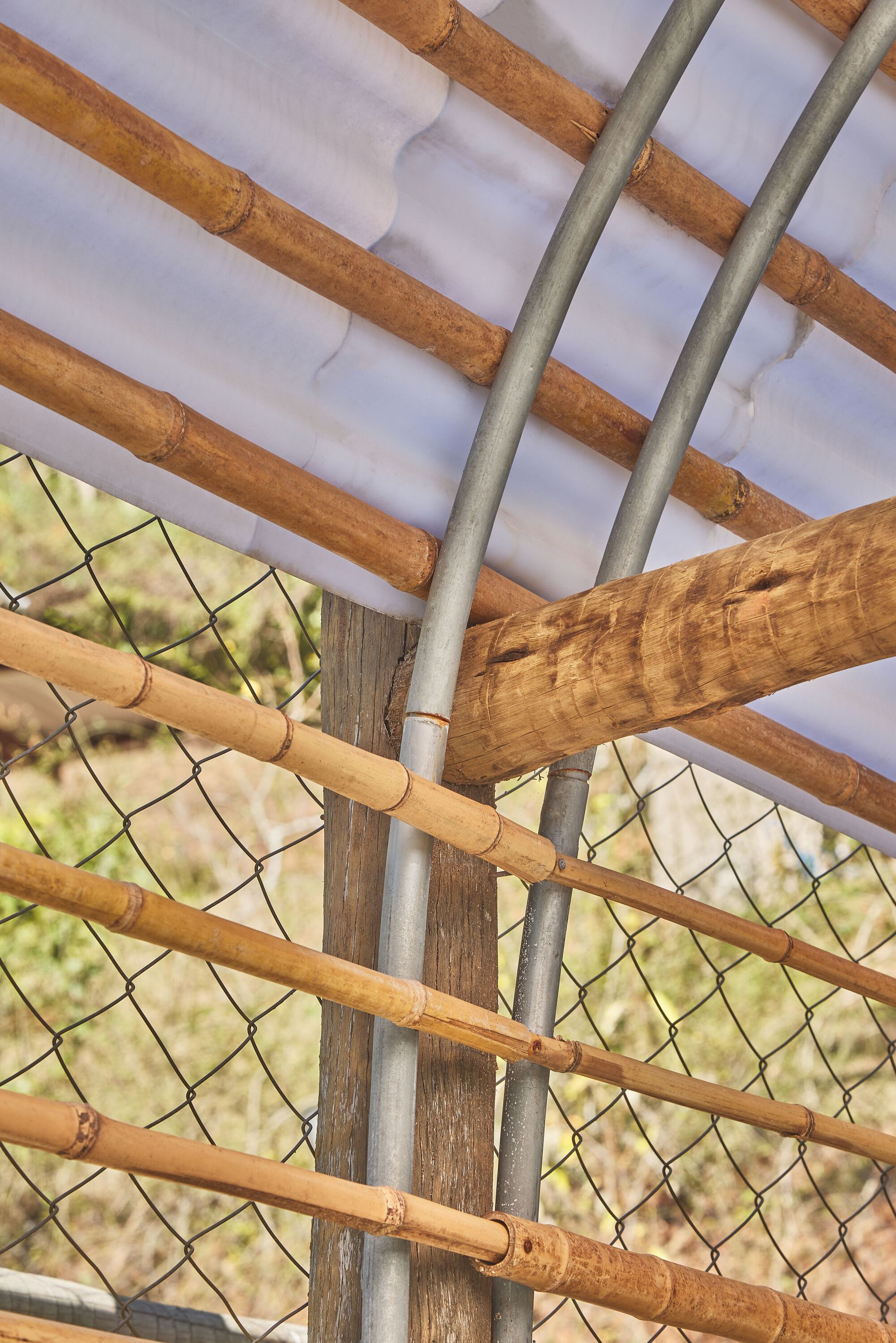 Detail of bamboo poles, chain-link mesh, and galvanized pipe junction at the pavilion corner