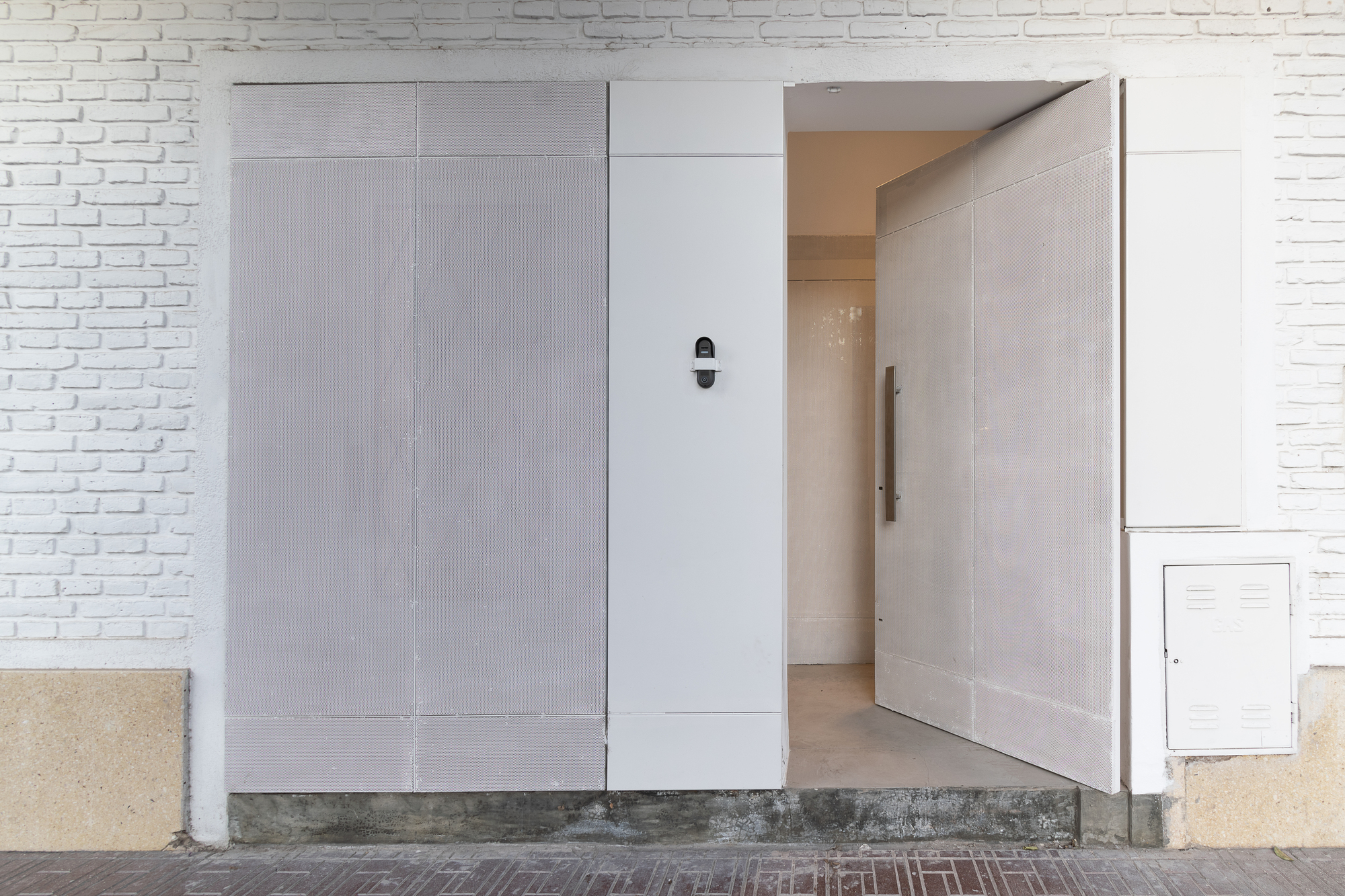 Street entry with pivoting grey panel door beside white painted brick and utility box