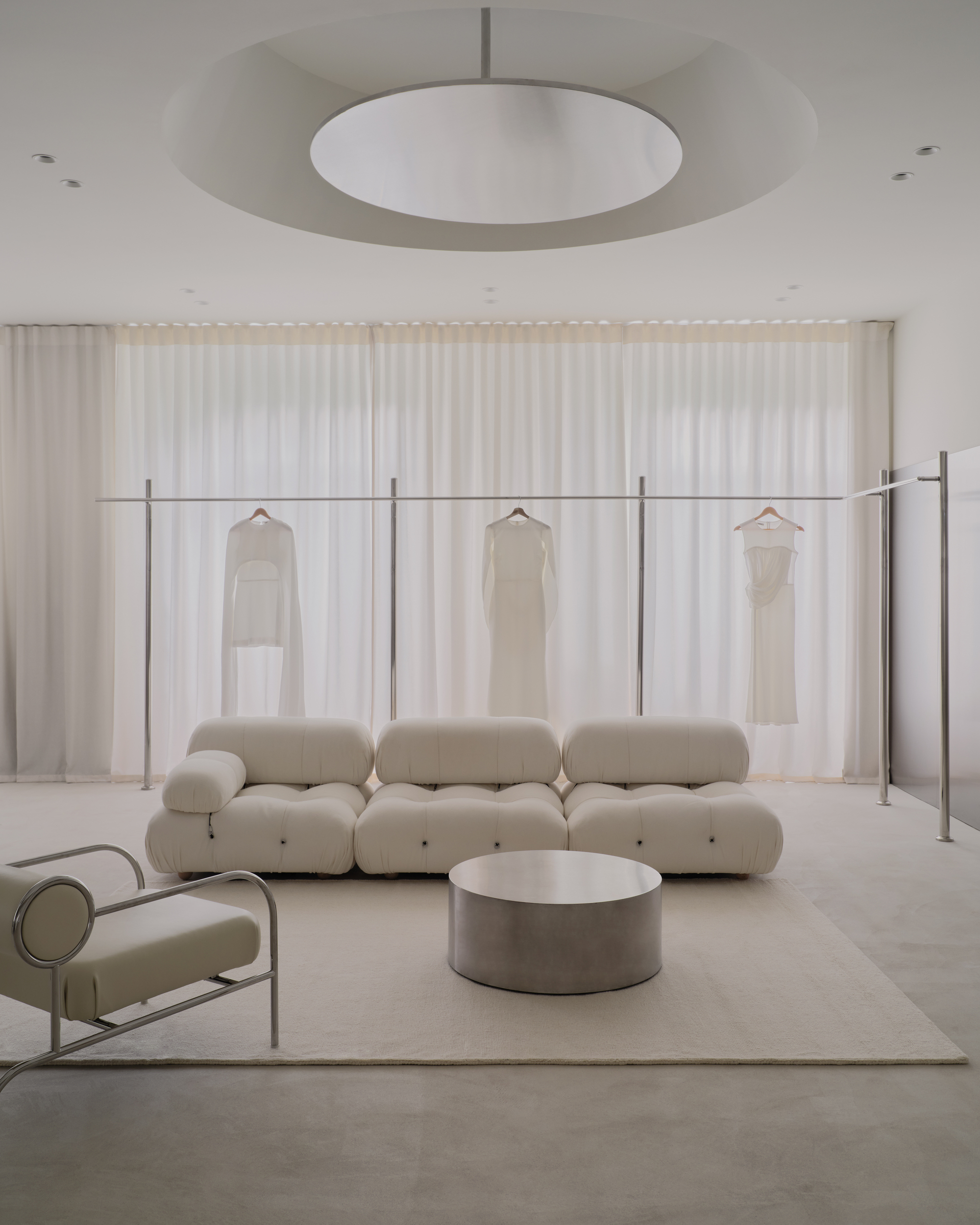 Freestanding steel clothing rack with hanging garments against sheer curtains and a disc pendant light