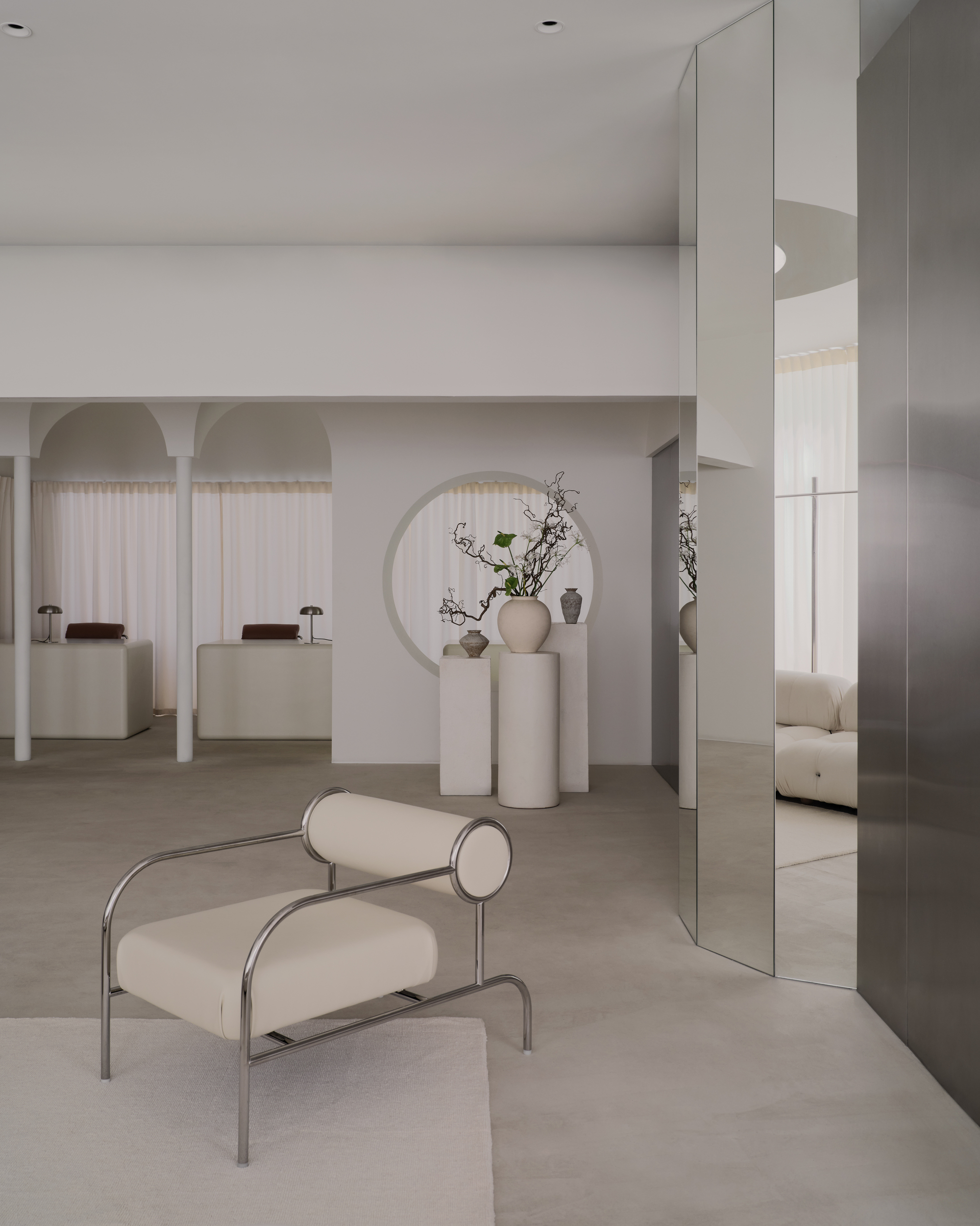 Reception area with tubular steel chair, mirrored panels, and cylindrical pedestals displaying ceramic vessels and branches