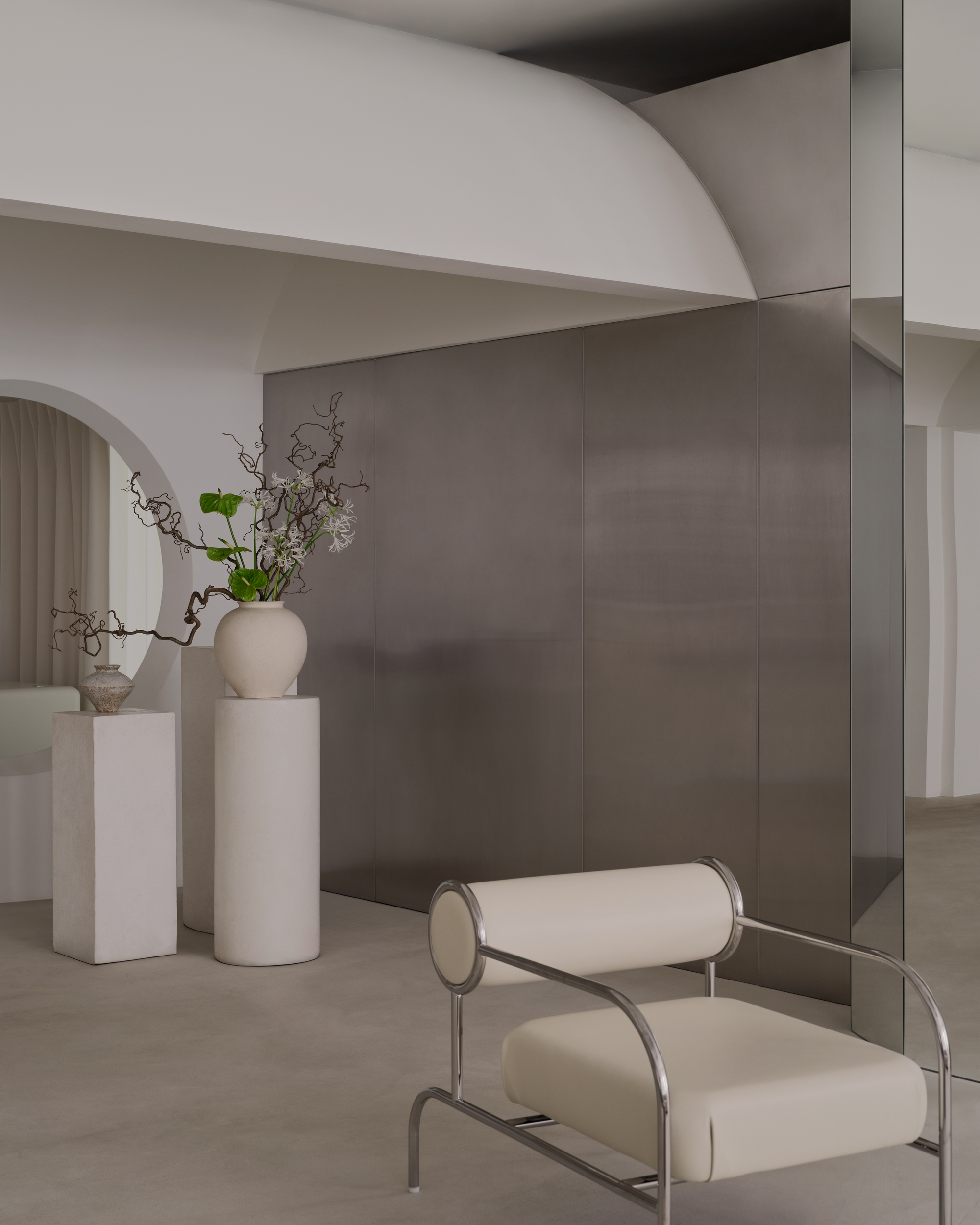 Close-up of cream upholstered chair with cylindrical armrests beside grey metal wall panels and white pedestals