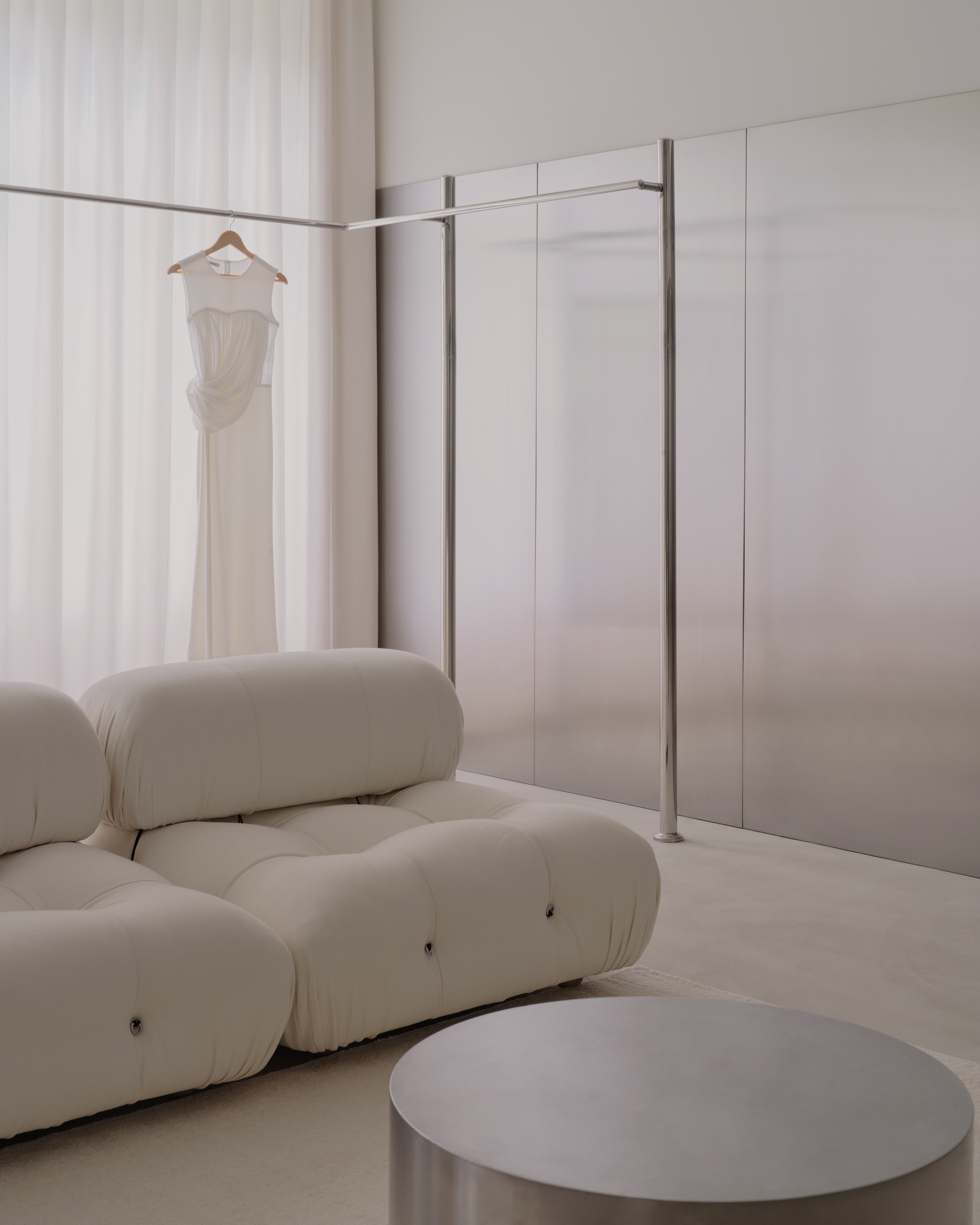 Dressing area with freestanding steel clothing rack, hanging white dress, and cushioned seating below sheer curtains