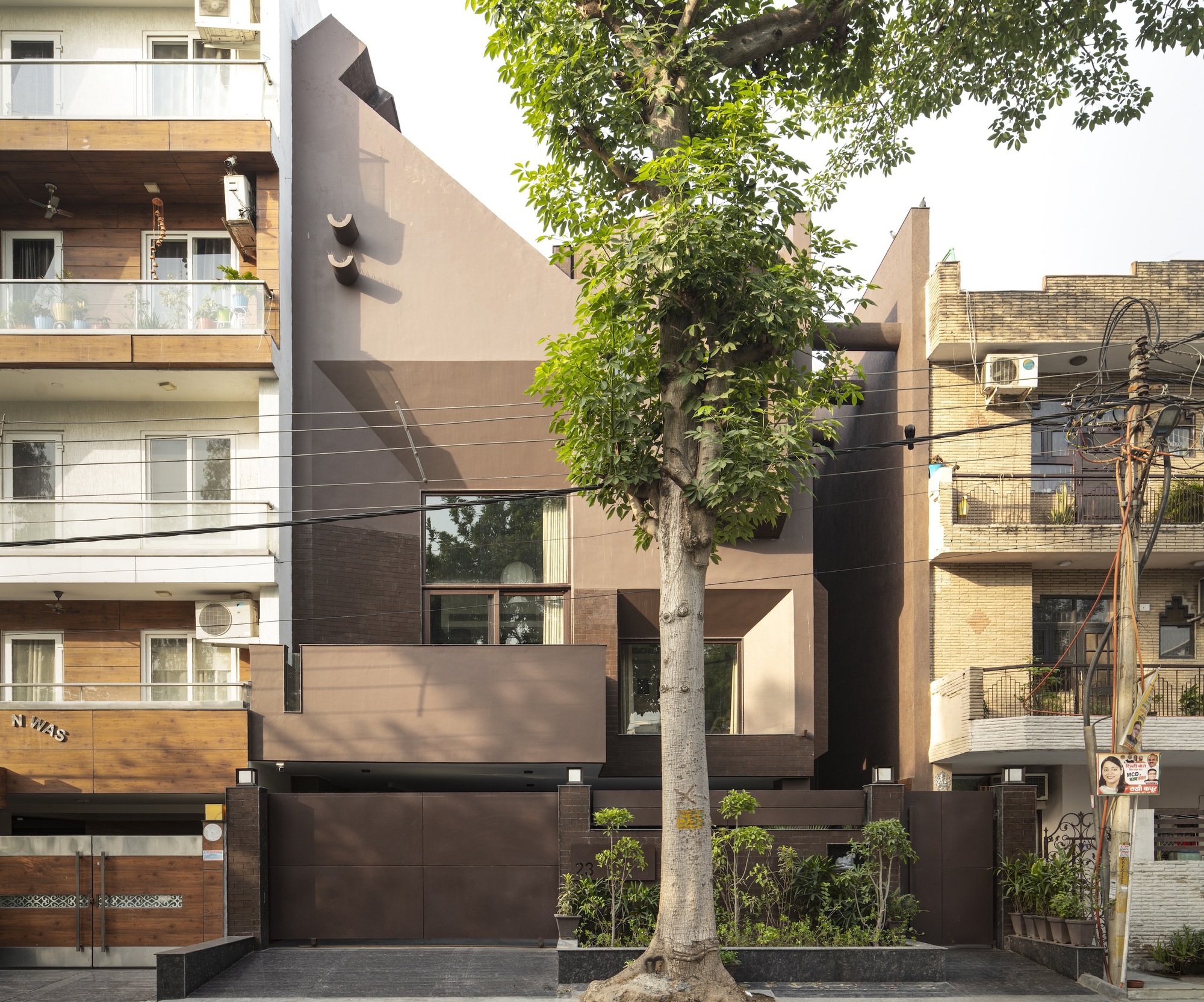 Street facade with layered brown and beige panels framed by a mature tree and neighboring buildings
