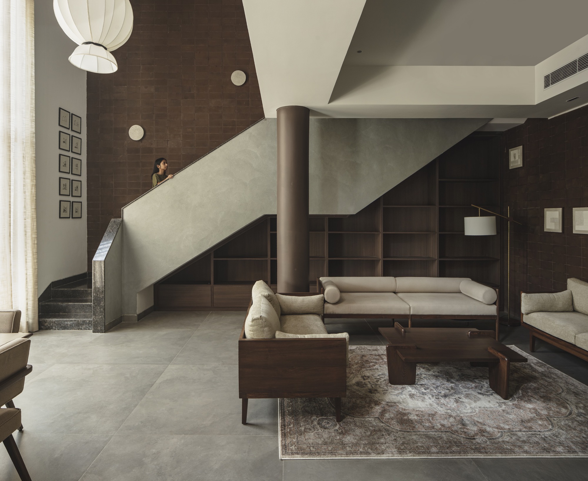 Double-height living space with pale staircase, cylindrical column, timber seating, and pendant light overhead