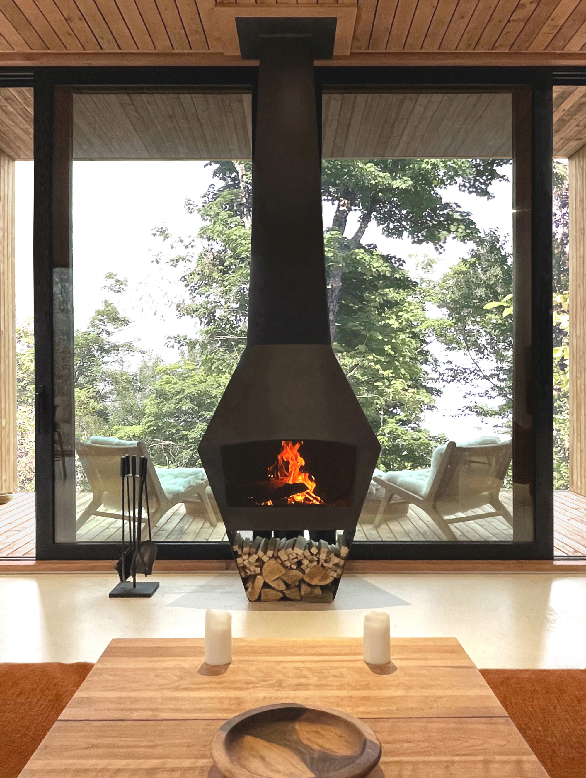 Freestanding fireplace with lit fire facing floor-to-ceiling windows overlooking trees and deck chairs