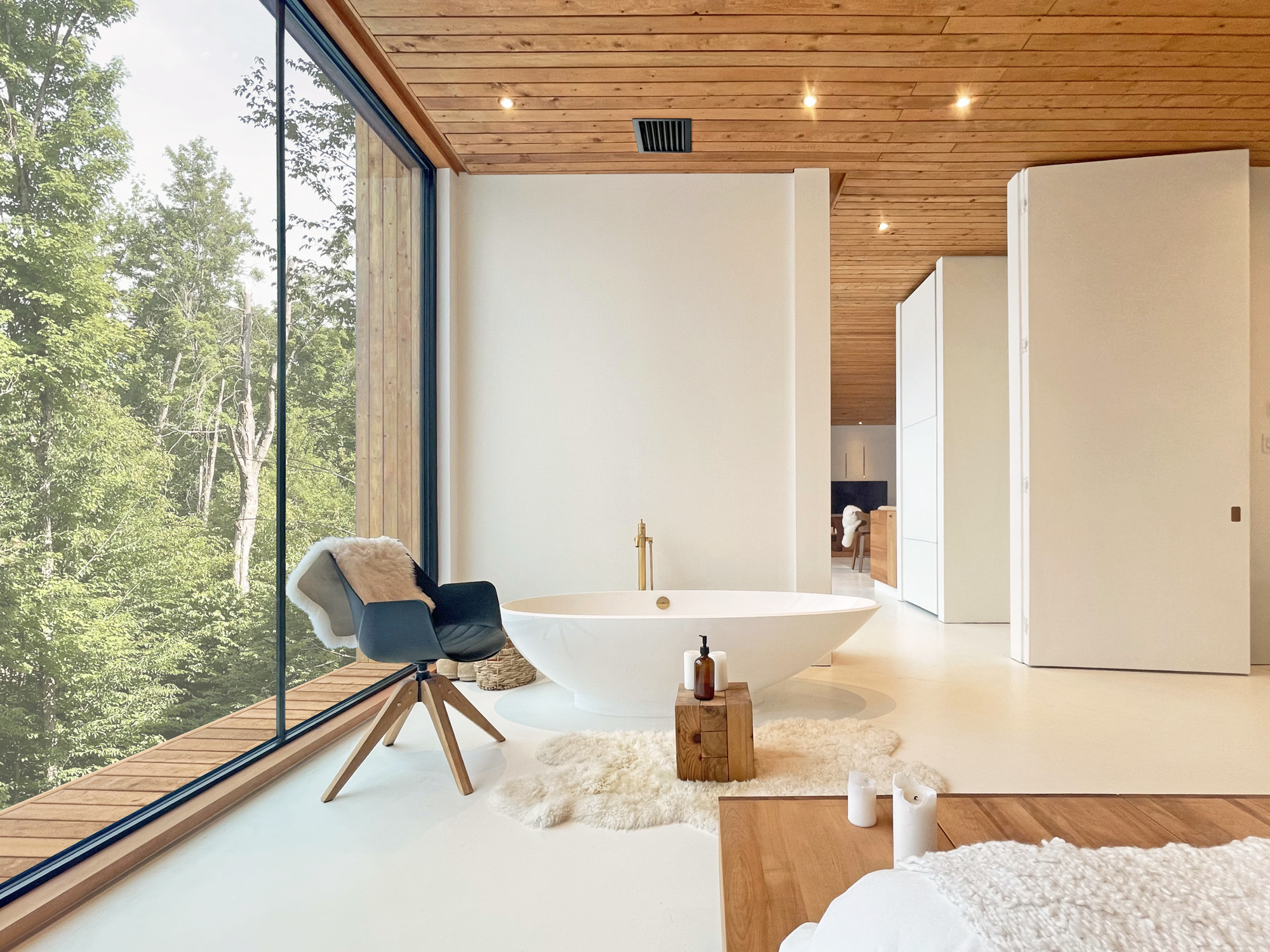 Freestanding bathtub on white flooring beneath timber ceiling with full-height glazing to forest beyond