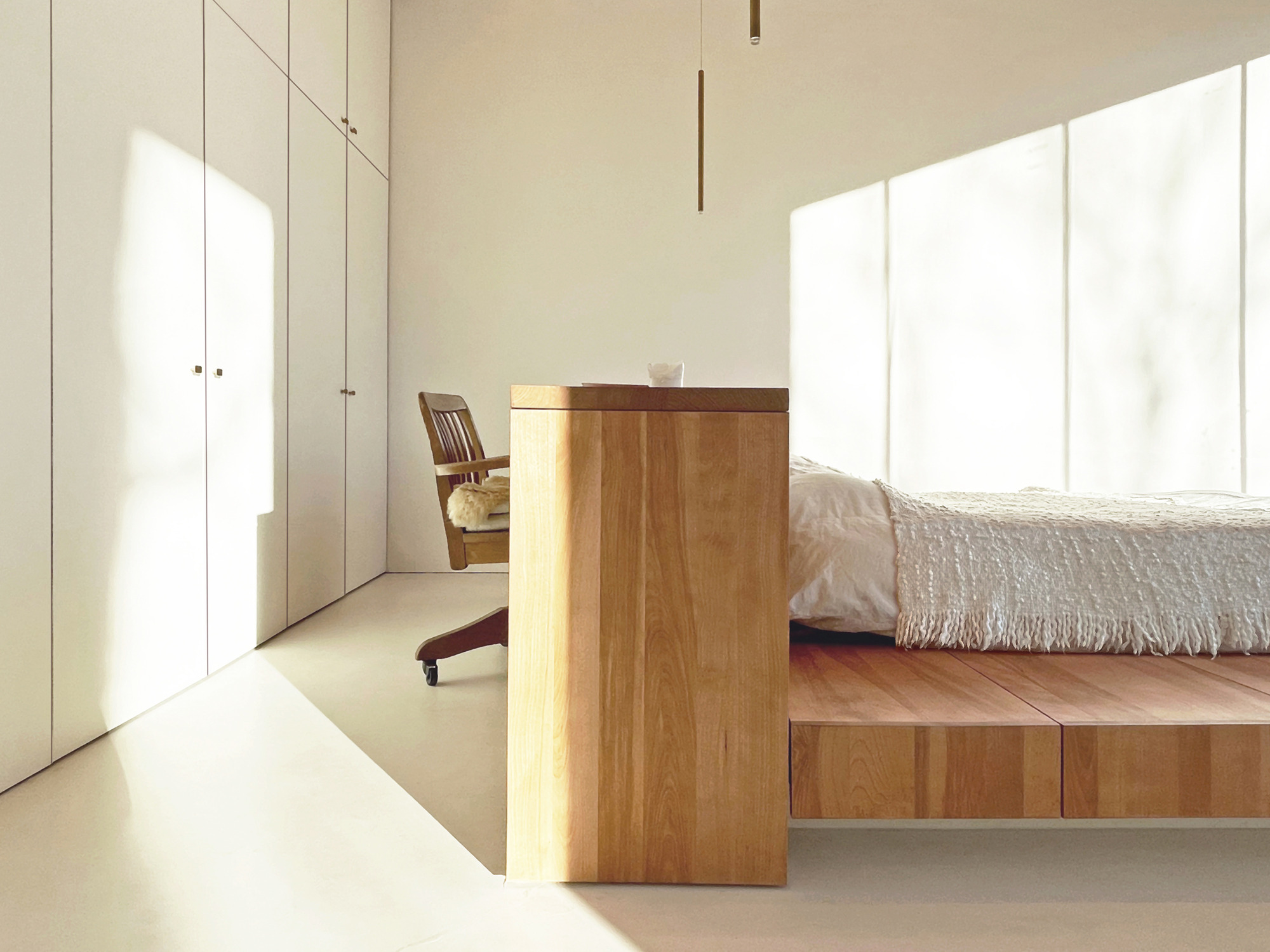 Minimalist bedroom with timber platform bed and floor-to-ceiling white storage cabinetry bathed in natural light