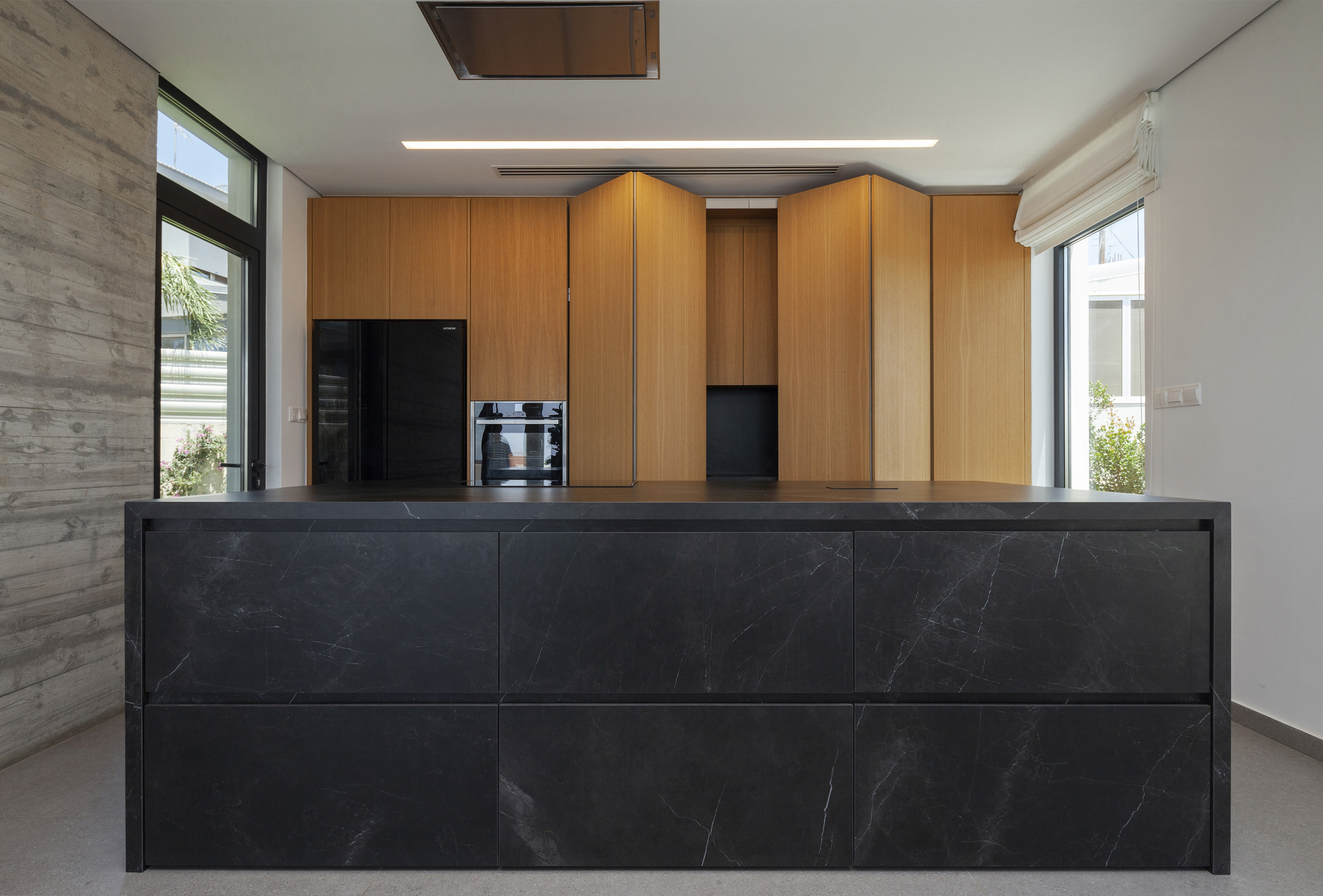 Kitchen island clad in dark marble with wood cabinetry beyond and flanking windows