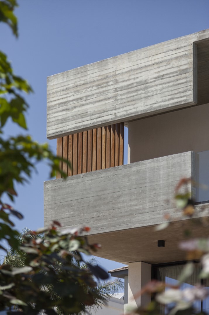 Stacked concrete volumes with vertical timber screening seen through foliage against a clear blue sky