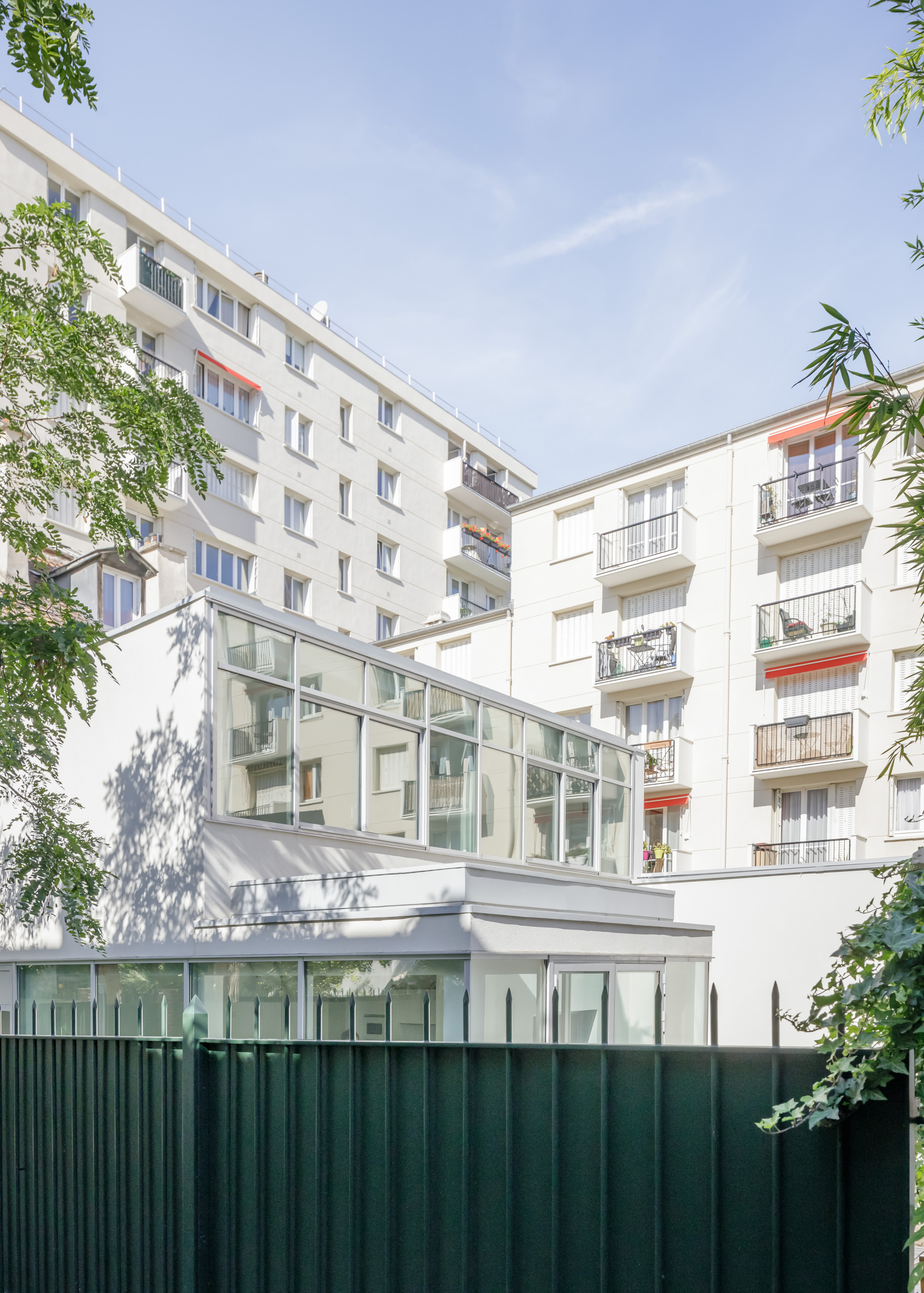 Glass pavilion with sloped skylight nestled between white residential buildings framed by foliage