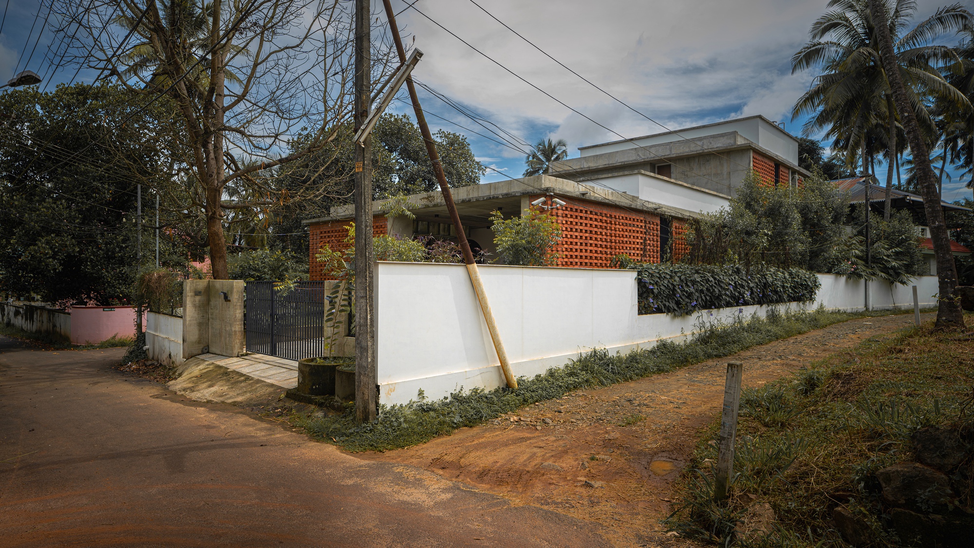 Street view showing the white compound wall, concrete roof, and perforated brick volumes among palm trees and utility poles