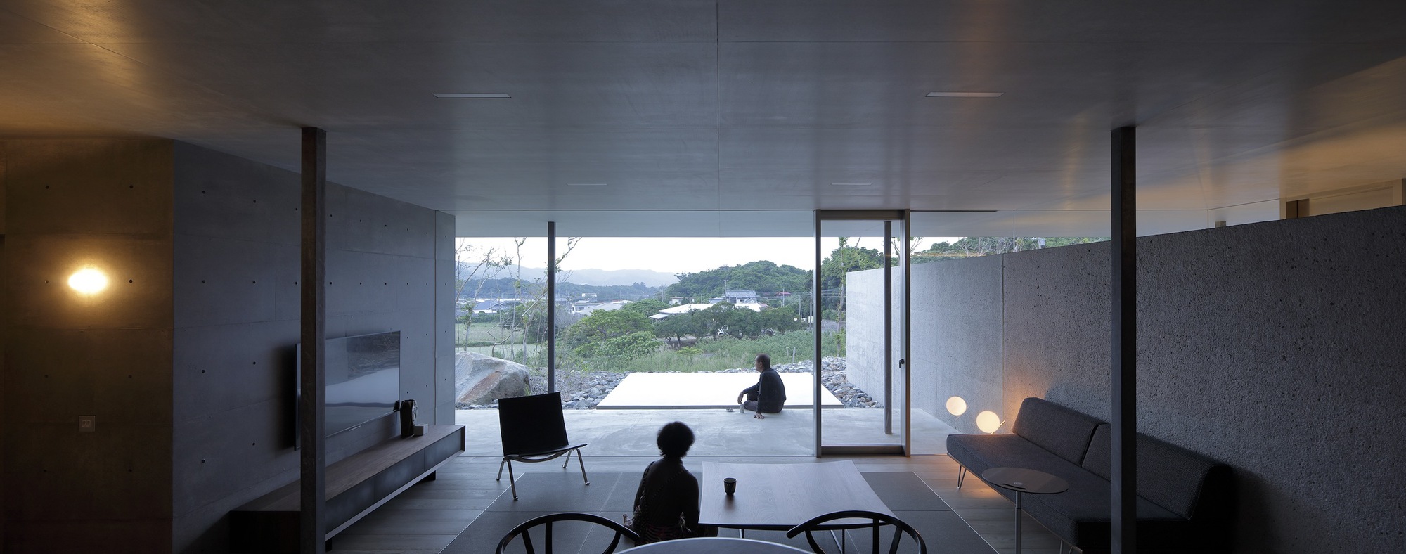 Living room interior at dusk with two figures seated and terrace opening to hillside beyond