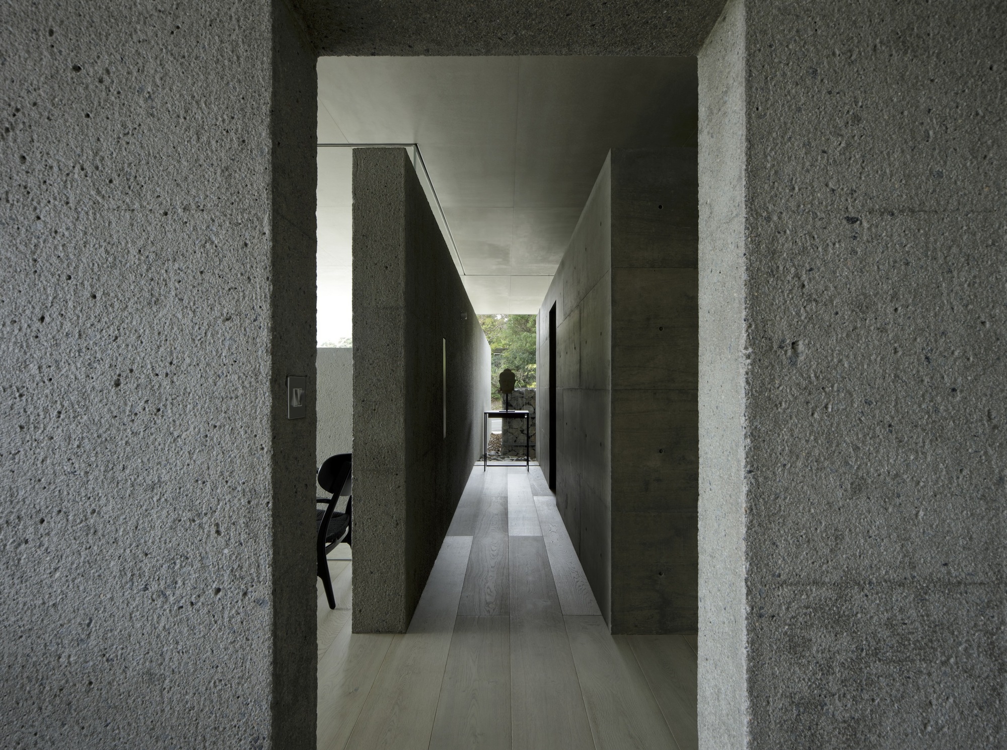 Corridor view through textured concrete walls leading to glazed opening with figure at table