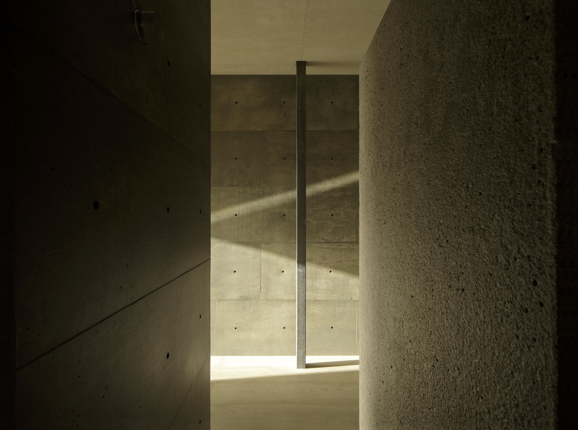 Narrow hallway in board-formed concrete with vertical slot window casting sunlight on textured stone wall