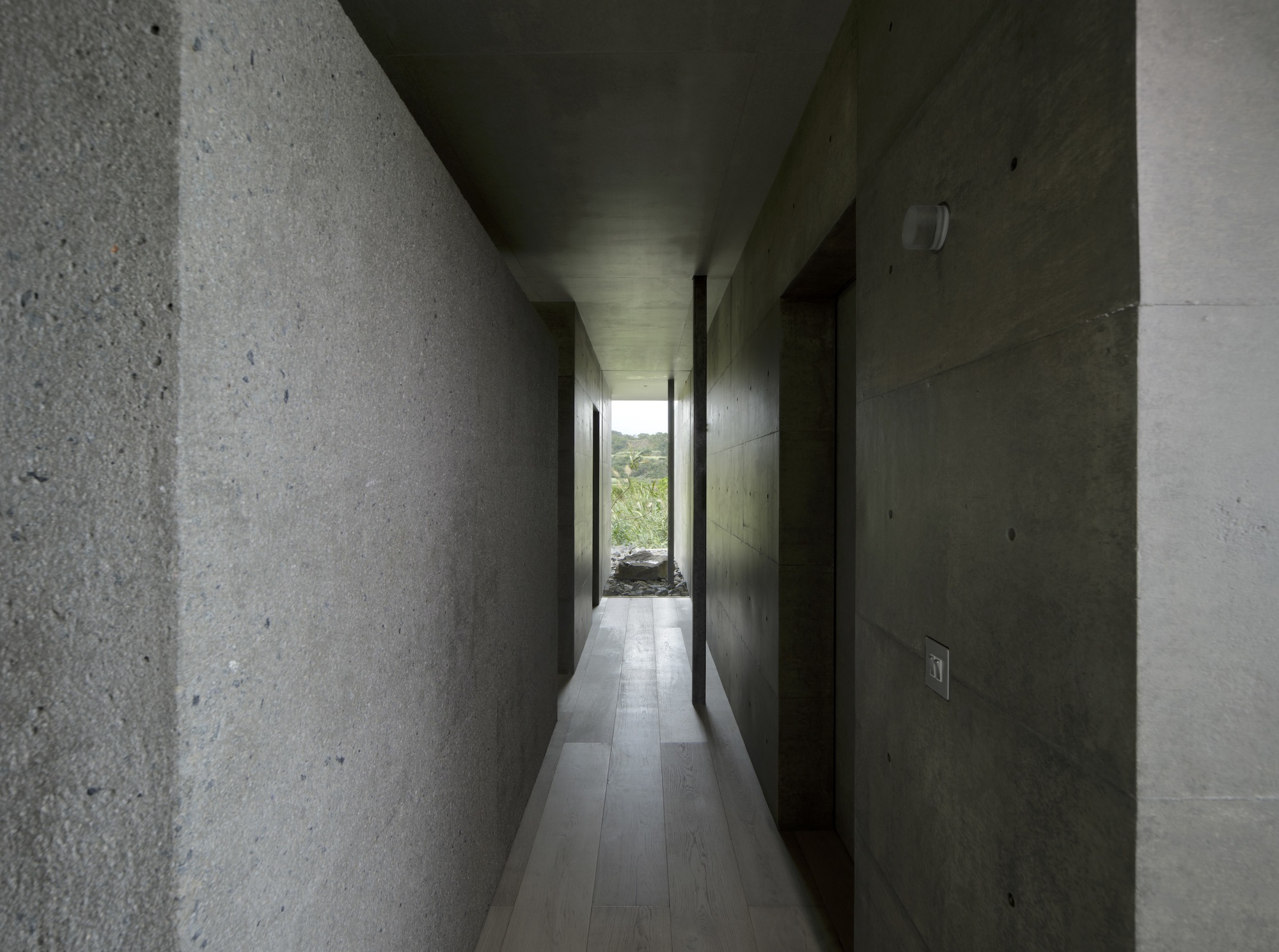 Long corridor lined with exposed concrete and rough stone walls leading to a sliver of garden