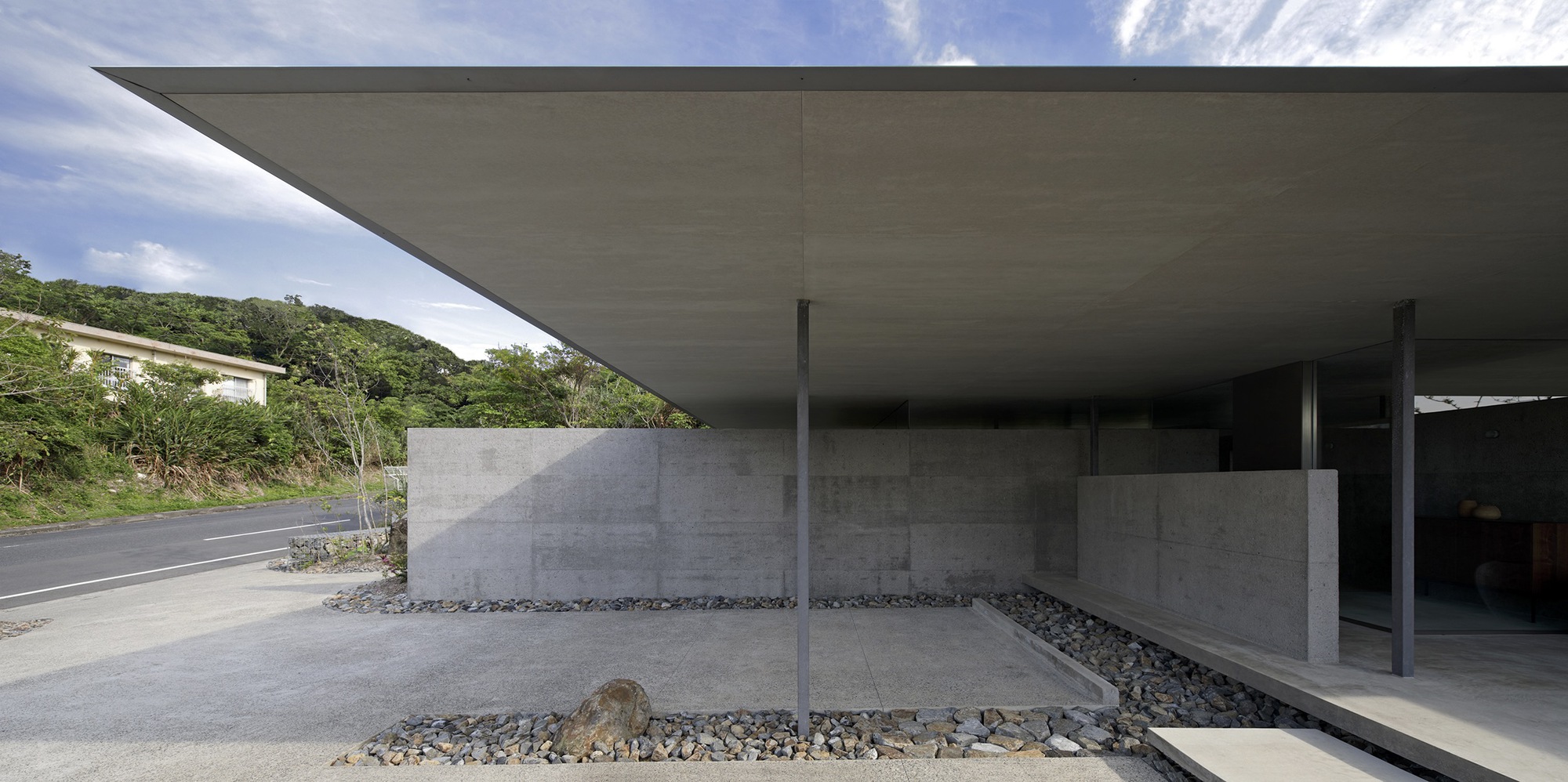 Covered entrance with thin steel columns and concrete walls set in a gravel courtyard beside a hillside road