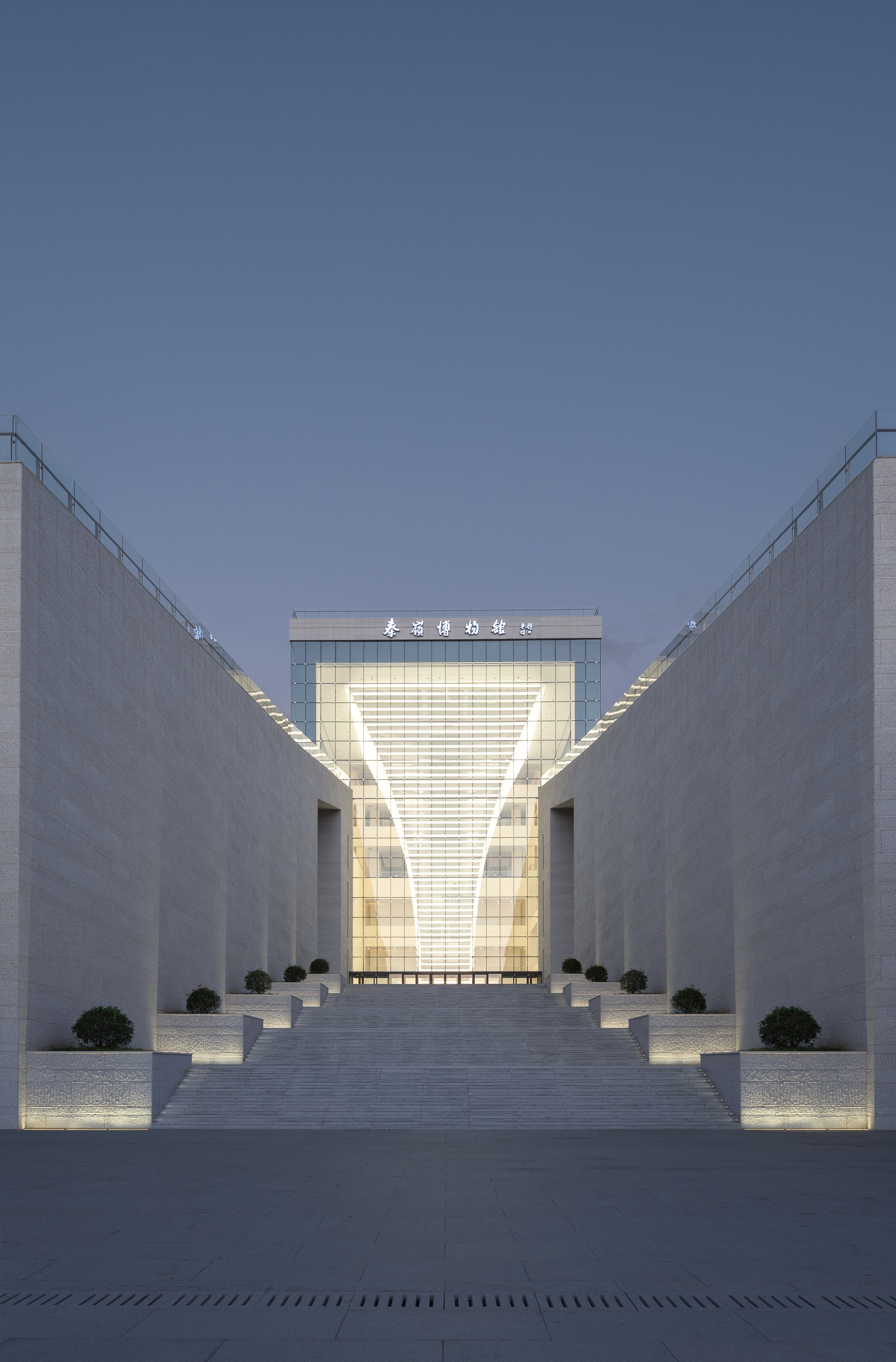 Stone-paved plaza flanked by angled walls leading to the illuminated glass entrance at dusk