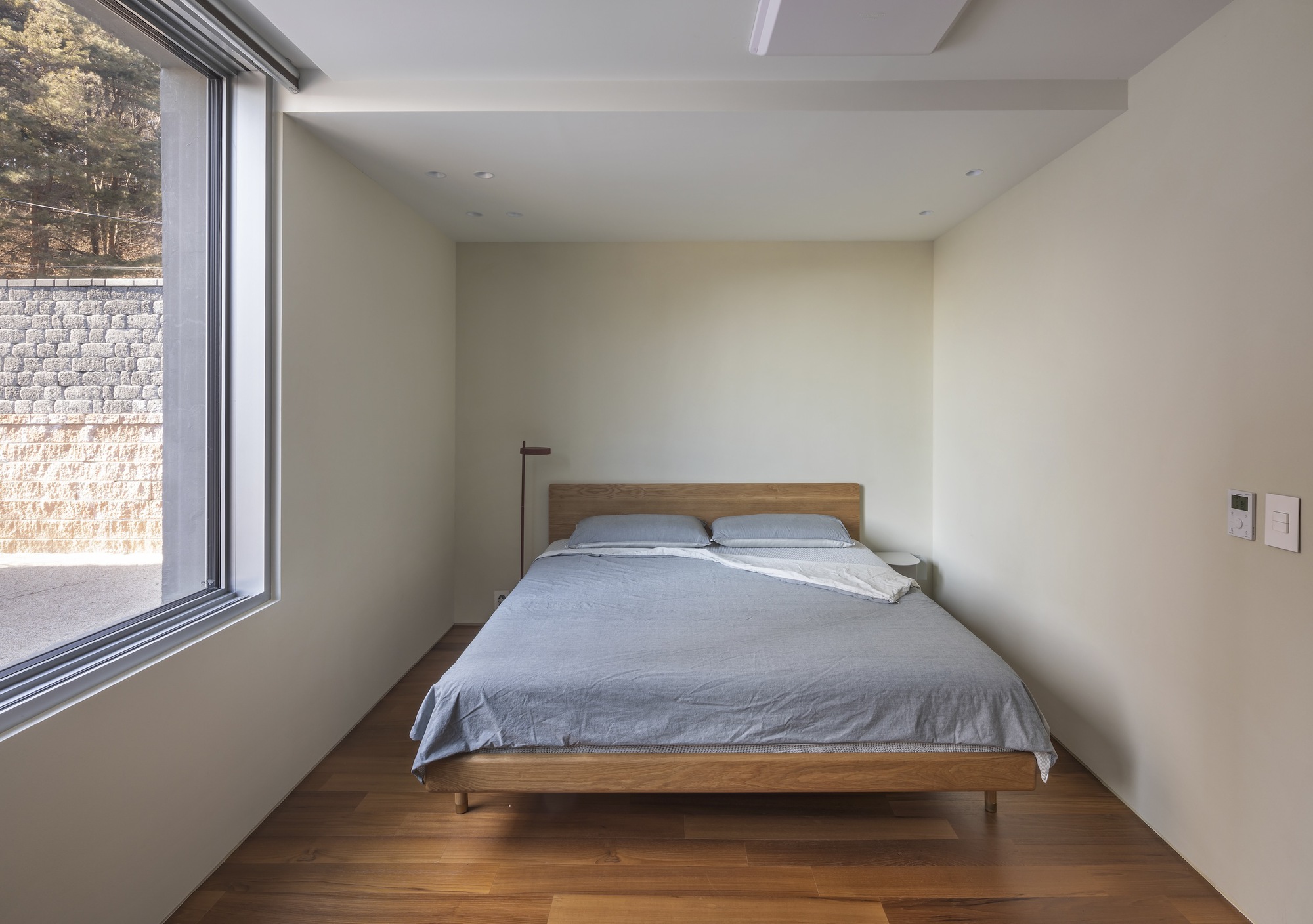 Bedroom with timber bed and wood floor beside full-height window overlooking stone retaining wall