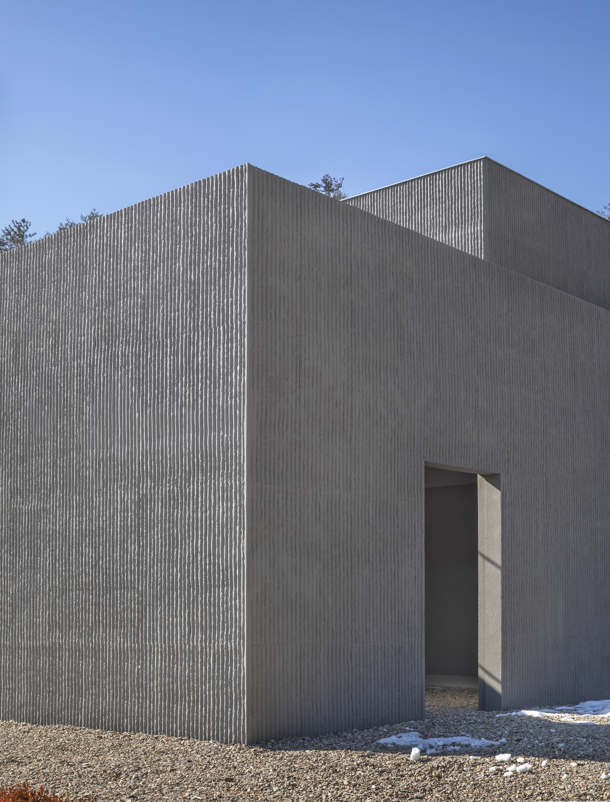 Exterior corner showing vertical concrete cladding with a narrow doorway and gravel ground under clear skies