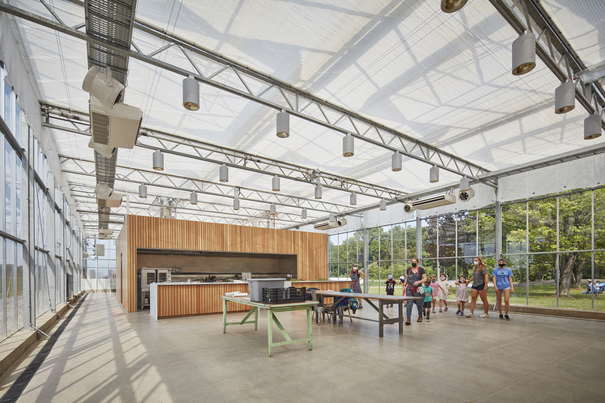 Interior gathering space with timber-clad kitchen module and ping-pong table under translucent roof panels and steel trusses