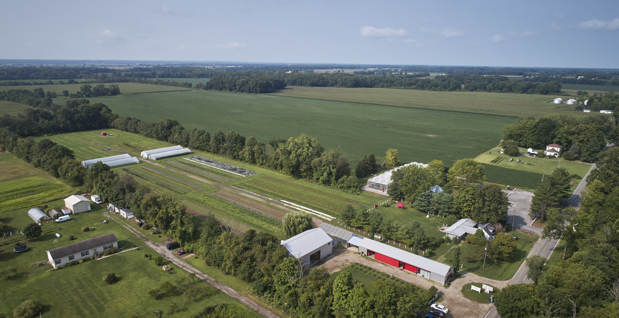 Aerial view of farm buildings with metal roofs and greenhouse structures set among cultivated fields and tree lines