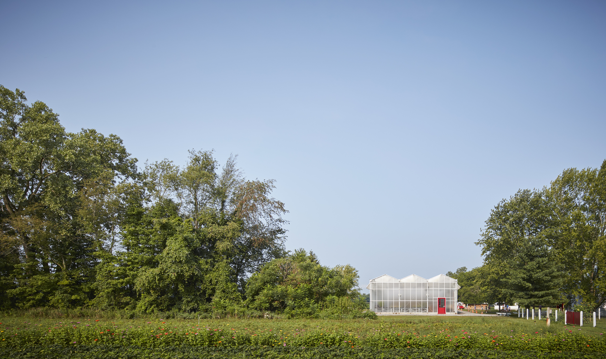 Greenhouse pavilion with translucent polycarbonate walls and red doors viewed across a green field beneath mature trees