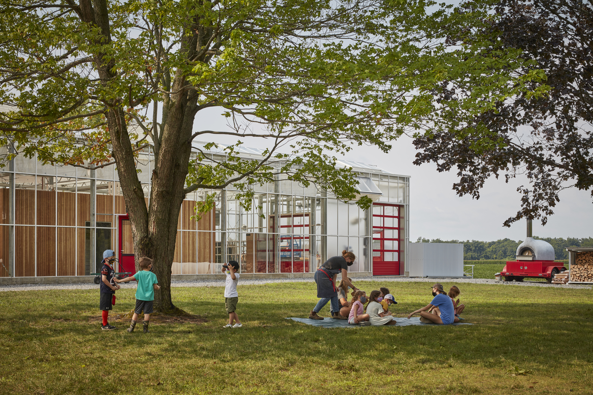 Children playing on the lawn in front of the greenhouse structure under the canopy of a large tree