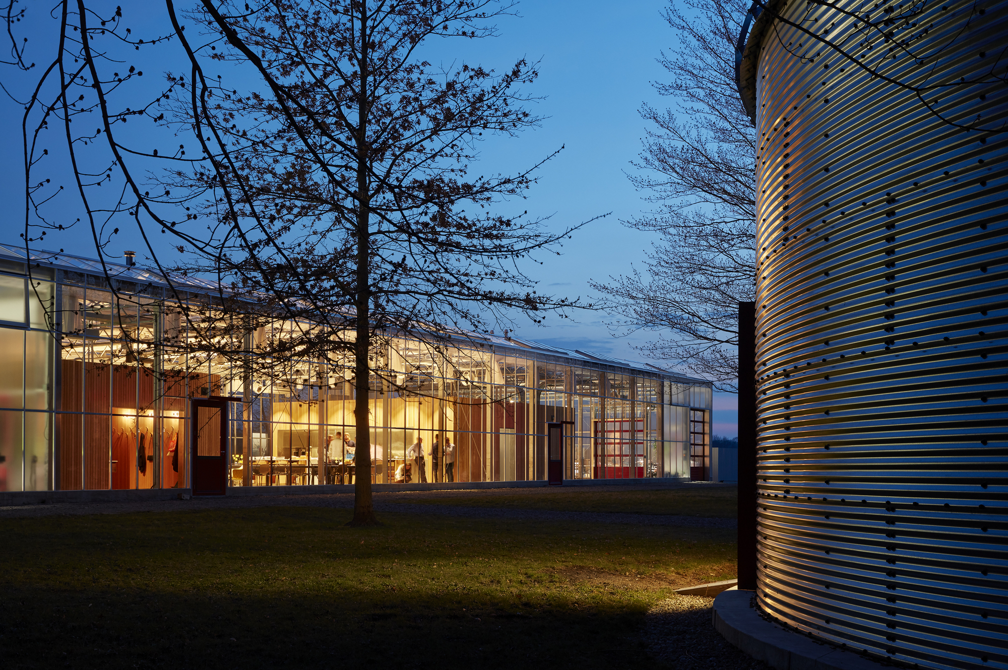 Glass-walled pavilion glowing from interior lights at dusk with bare trees and a cylindrical metal form nearby
