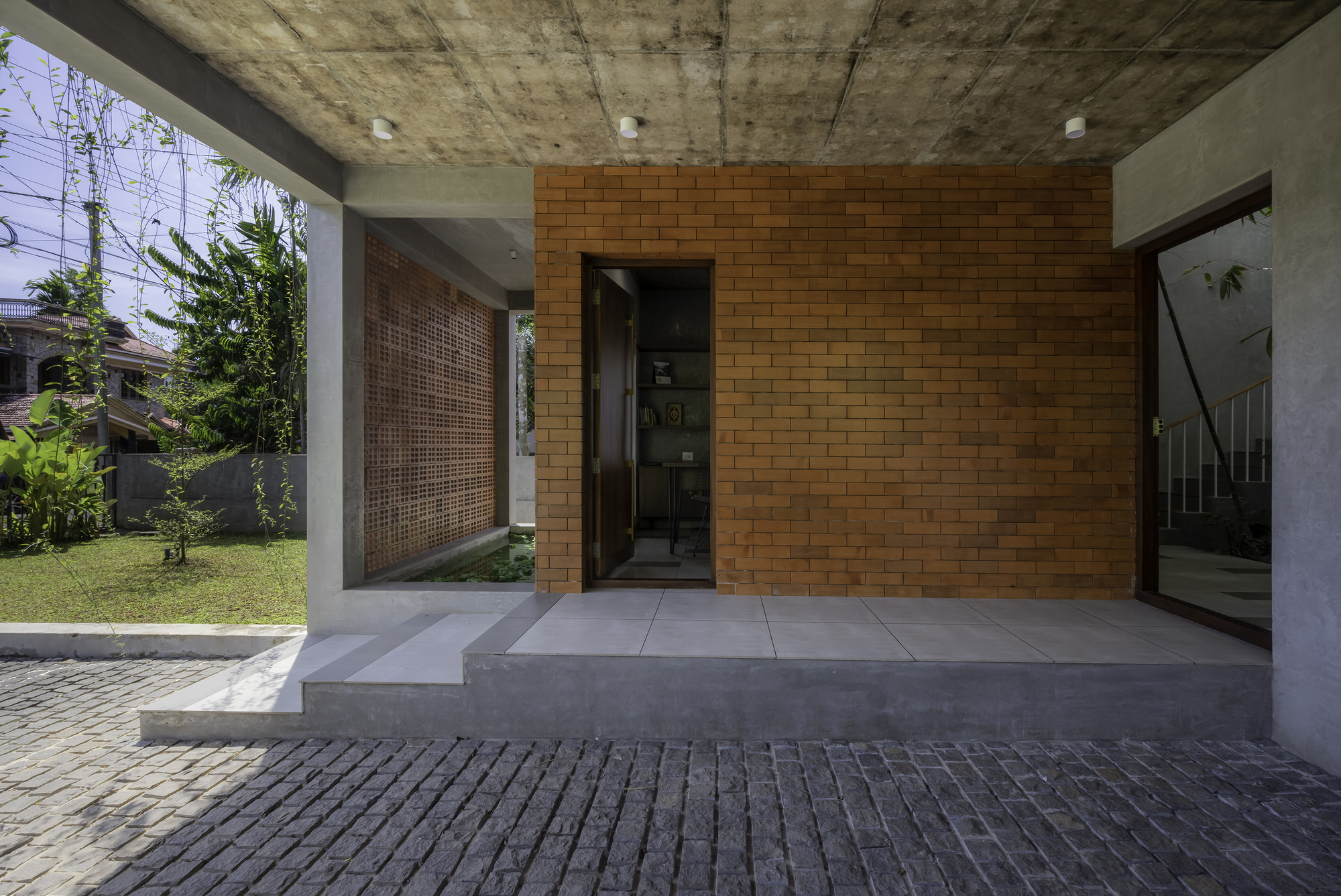 Covered entry terrace with exposed concrete ceiling, orange brick wall and planted courtyard beyond