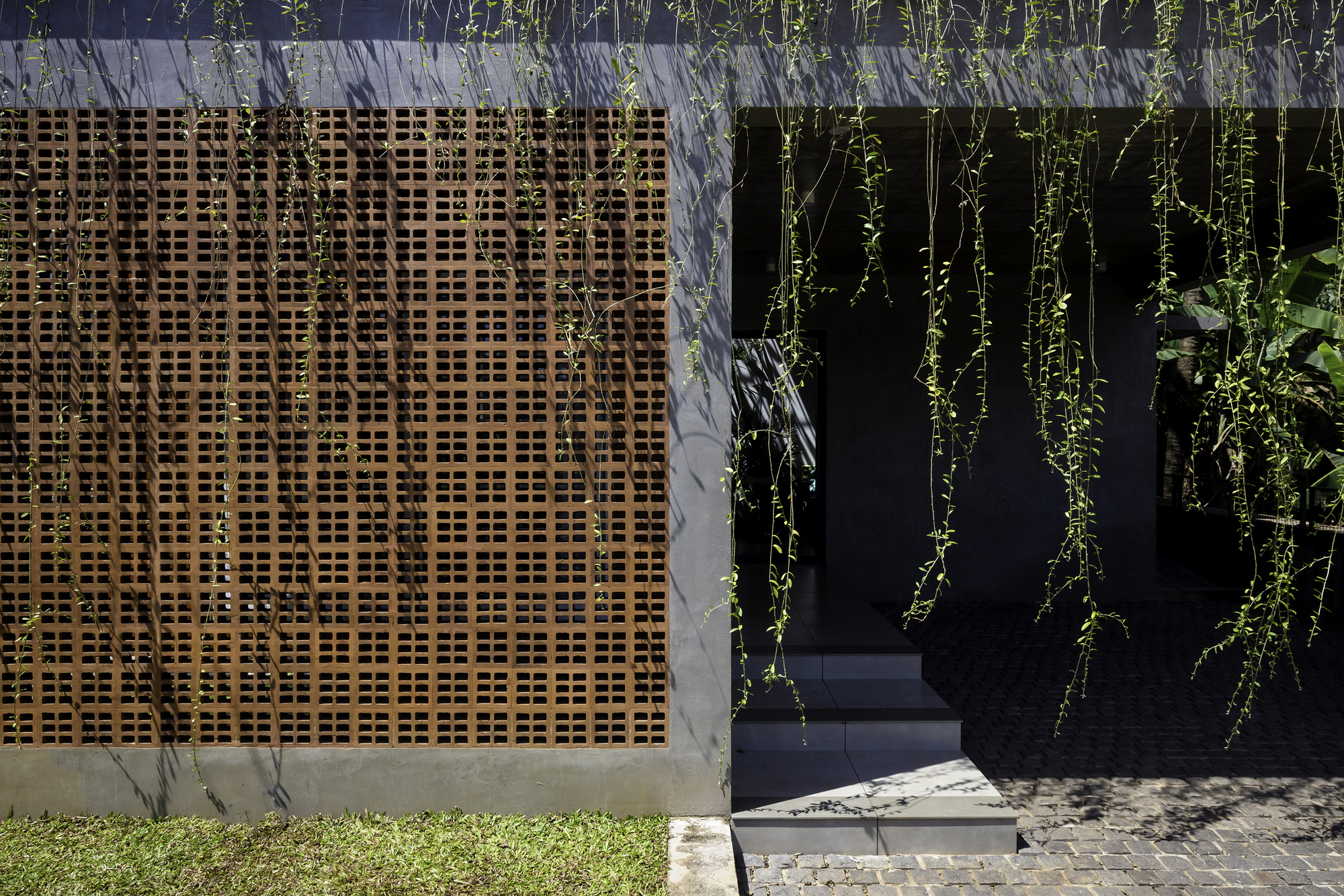 Perforated timber screen beside a dark entrance framed by cascading greenery and lawn edge