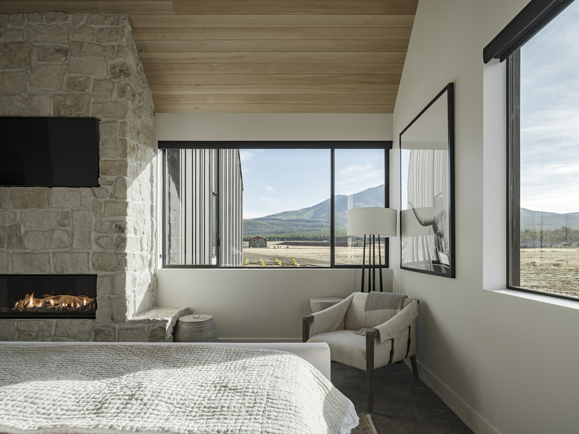 Bedroom with corner windows framing mountain views and stone fireplace with timber ceiling