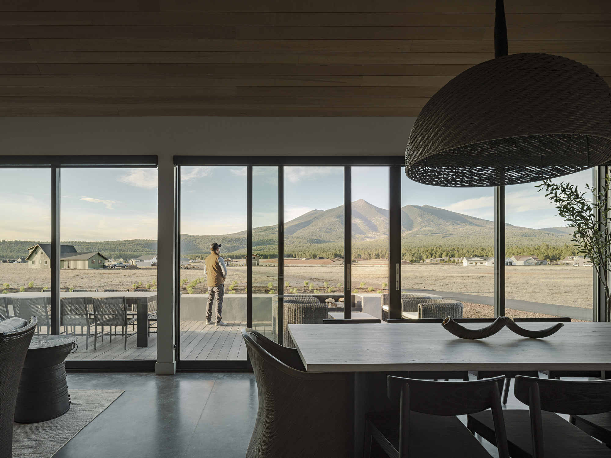 Dining area with full-height glazing and a figure standing on the terrace framed by distant peaks