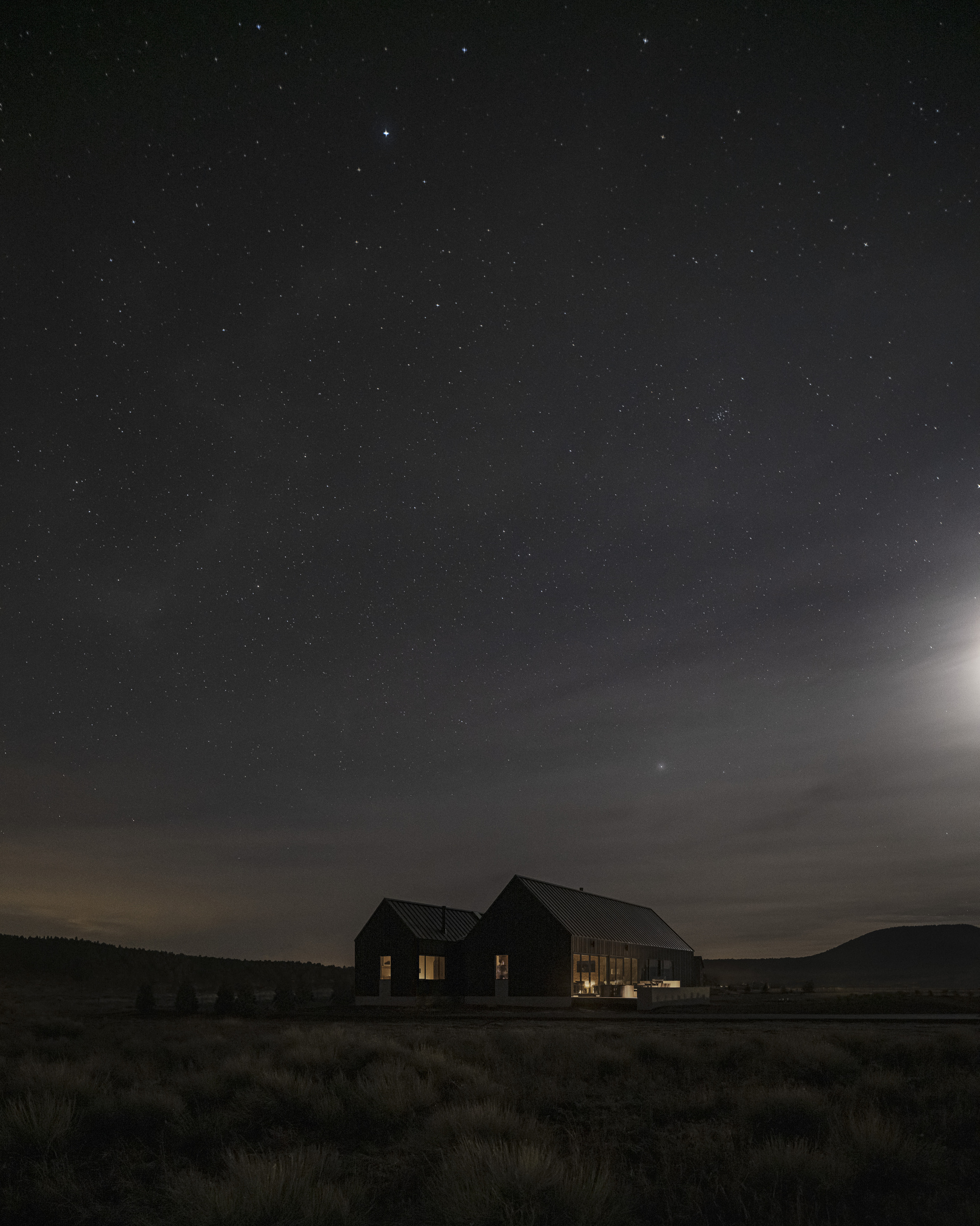 Twin gabled volumes illuminated from within across a scrubland field under a star-filled night sky