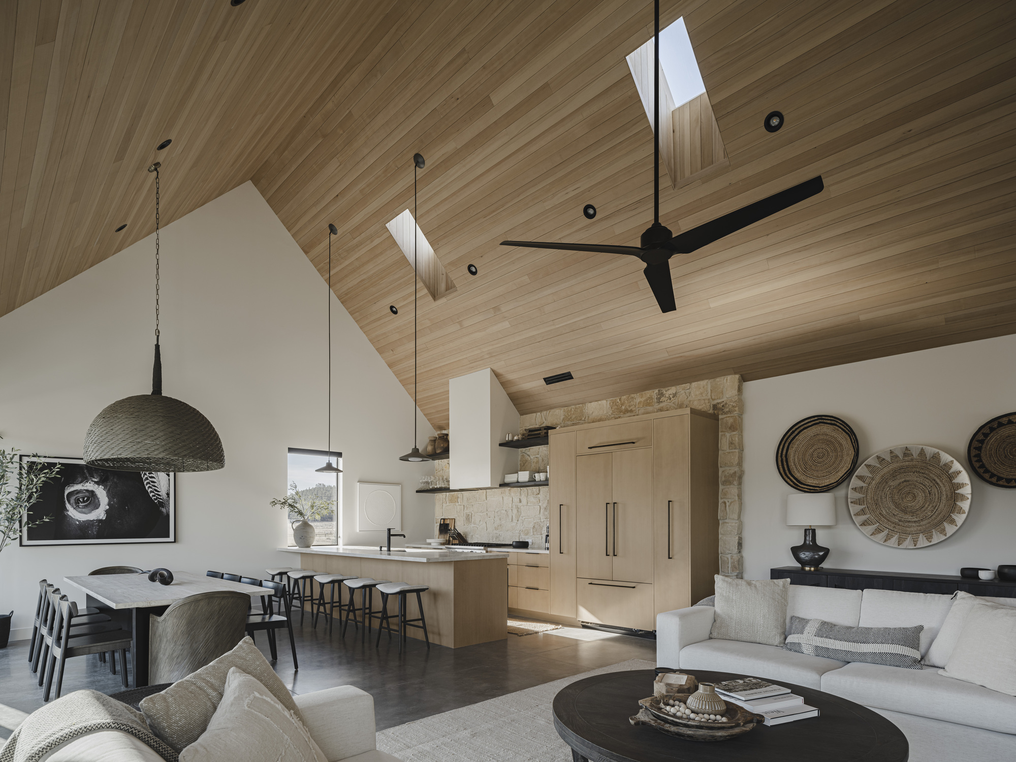 Open living space with vaulted wood ceiling, skylights, stone fireplace wall and pendant lighting over seating